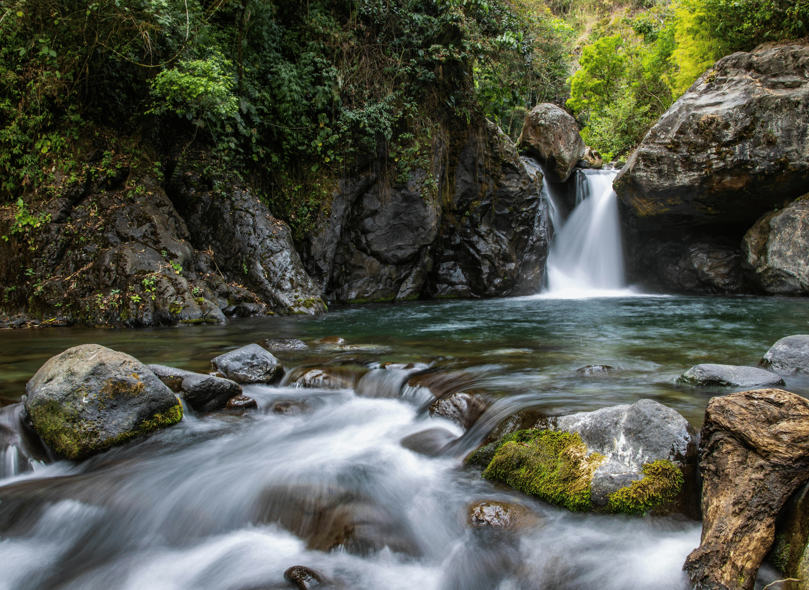 A flowing waterfall cascading over mossy rocks into a small pool in a shaded, rocky setting.