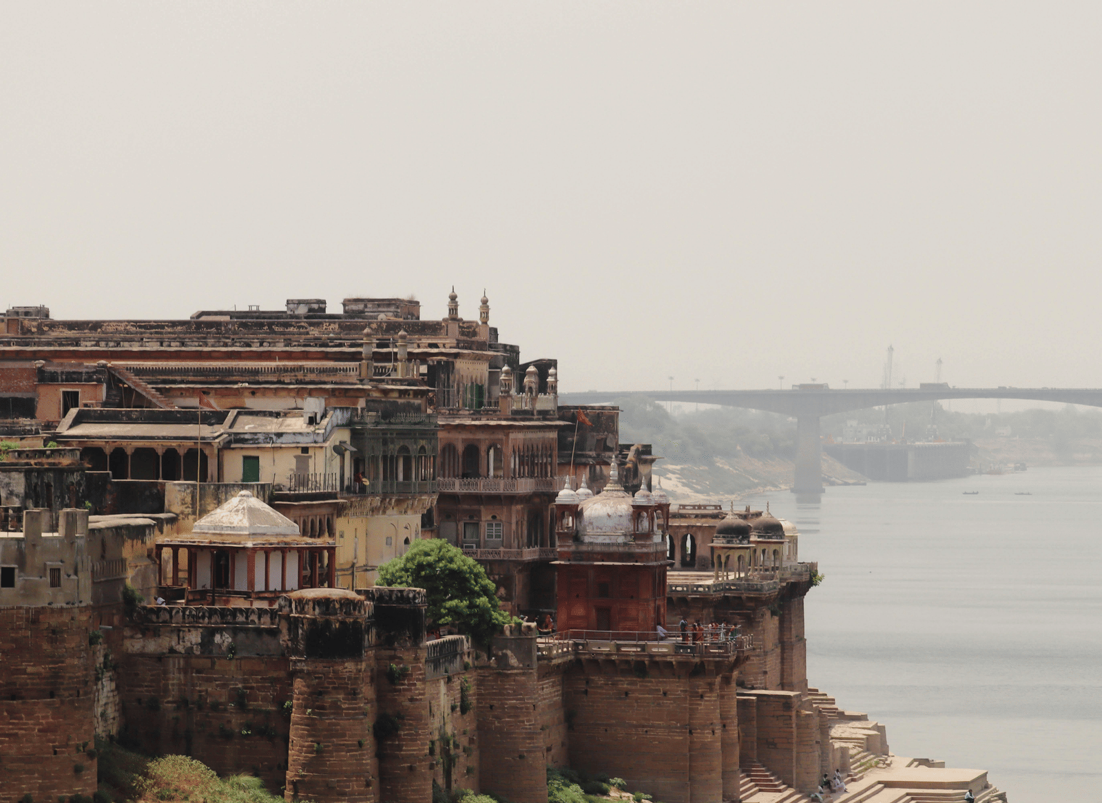An ancient, tiered city structure of red stone besides the bank of a river under a hazy sky.