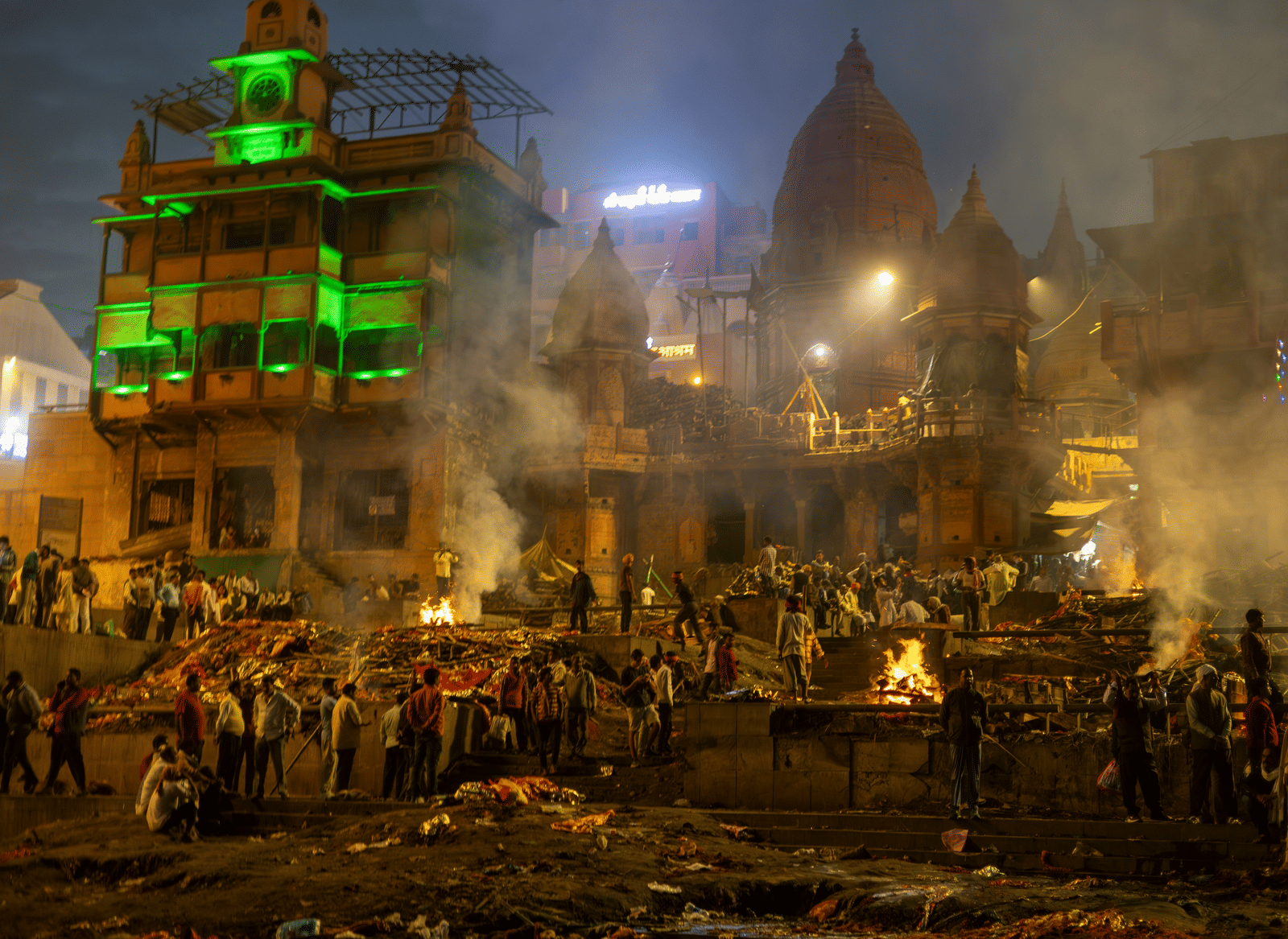 A somber view of a cremation ceremony taking place on a riverside fort at night, illuminated by firelight.