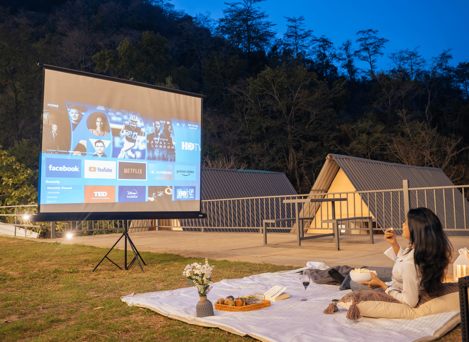 A person sits on a picnic blanket watching a film projected onto a screen at Perfect Stayz The Jungle Resort.