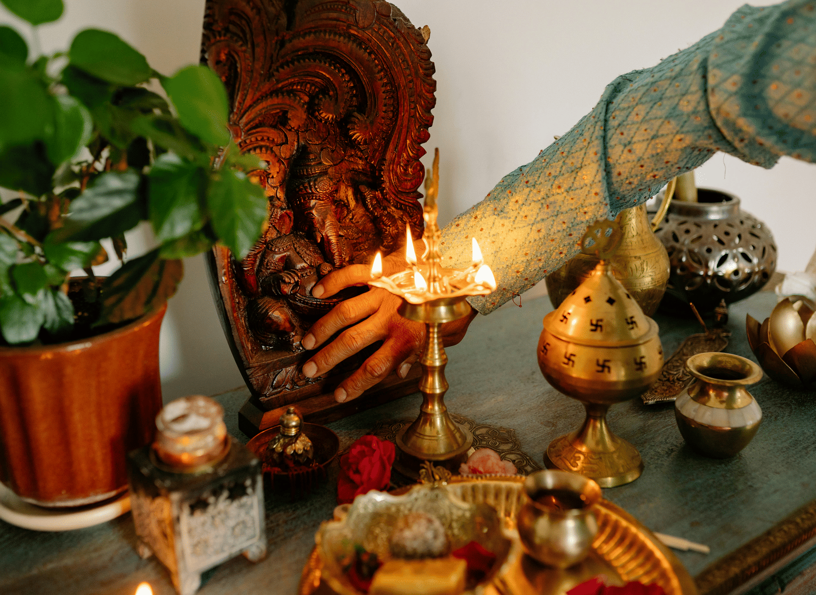 A lit lamp sits on a table with offerings and a plant.