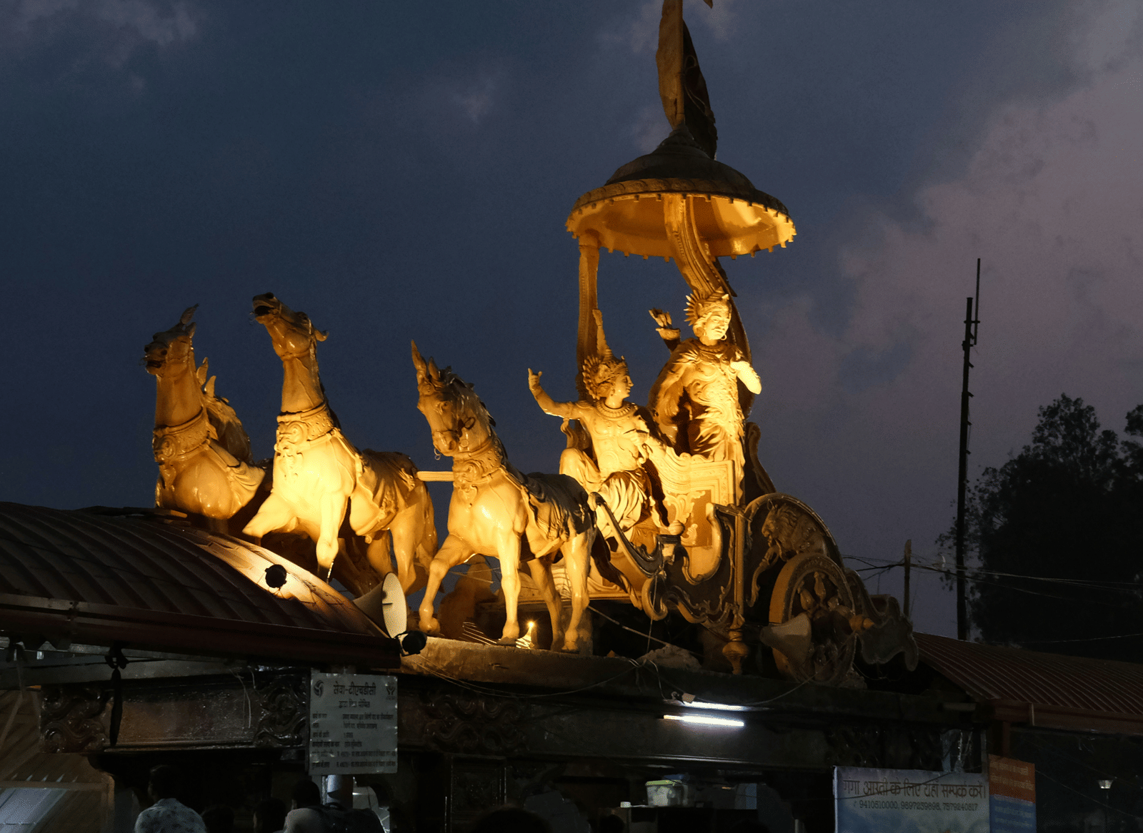 A brightly illuminated golden chariot statue, depicting Lord Krishna and Arjun against the night sky.