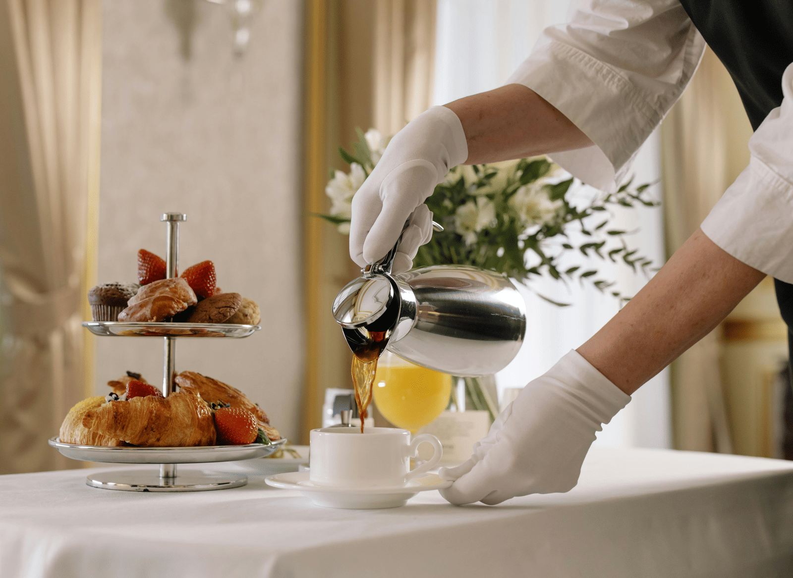 A person wearing white gloves pours tea into a cup on a table set with a vase, pastries, fruit, and juice.