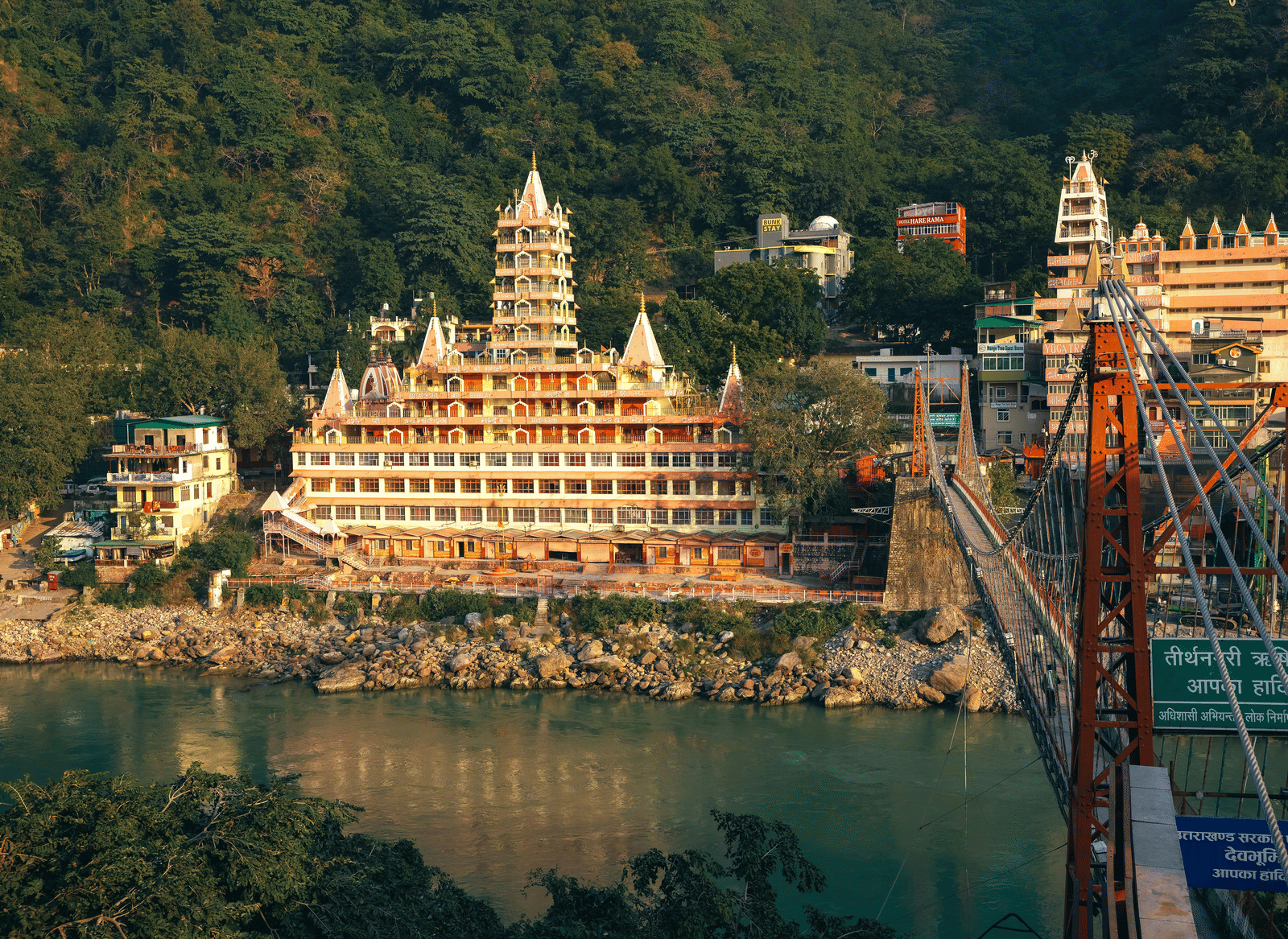 A suspension bridge across a river, with a large multi-storied building, and forested hills in the background.