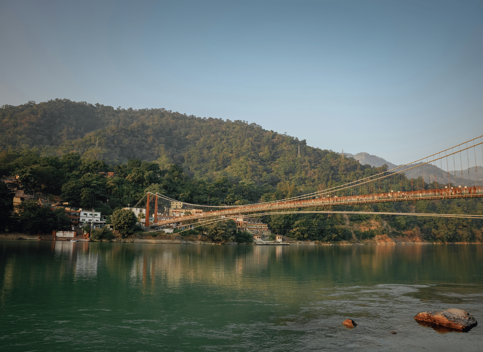A suspension bridge over a river, with forested hills in the background, under a bright sky.