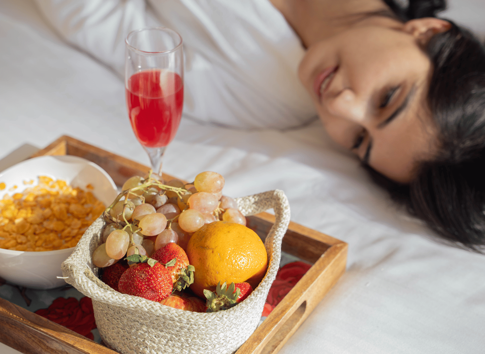 A woman in white robes lying beside a tray of fruits, a yellow dish, and a glass filled with red wine | Perfectstayz Value Hills, Rishikesh