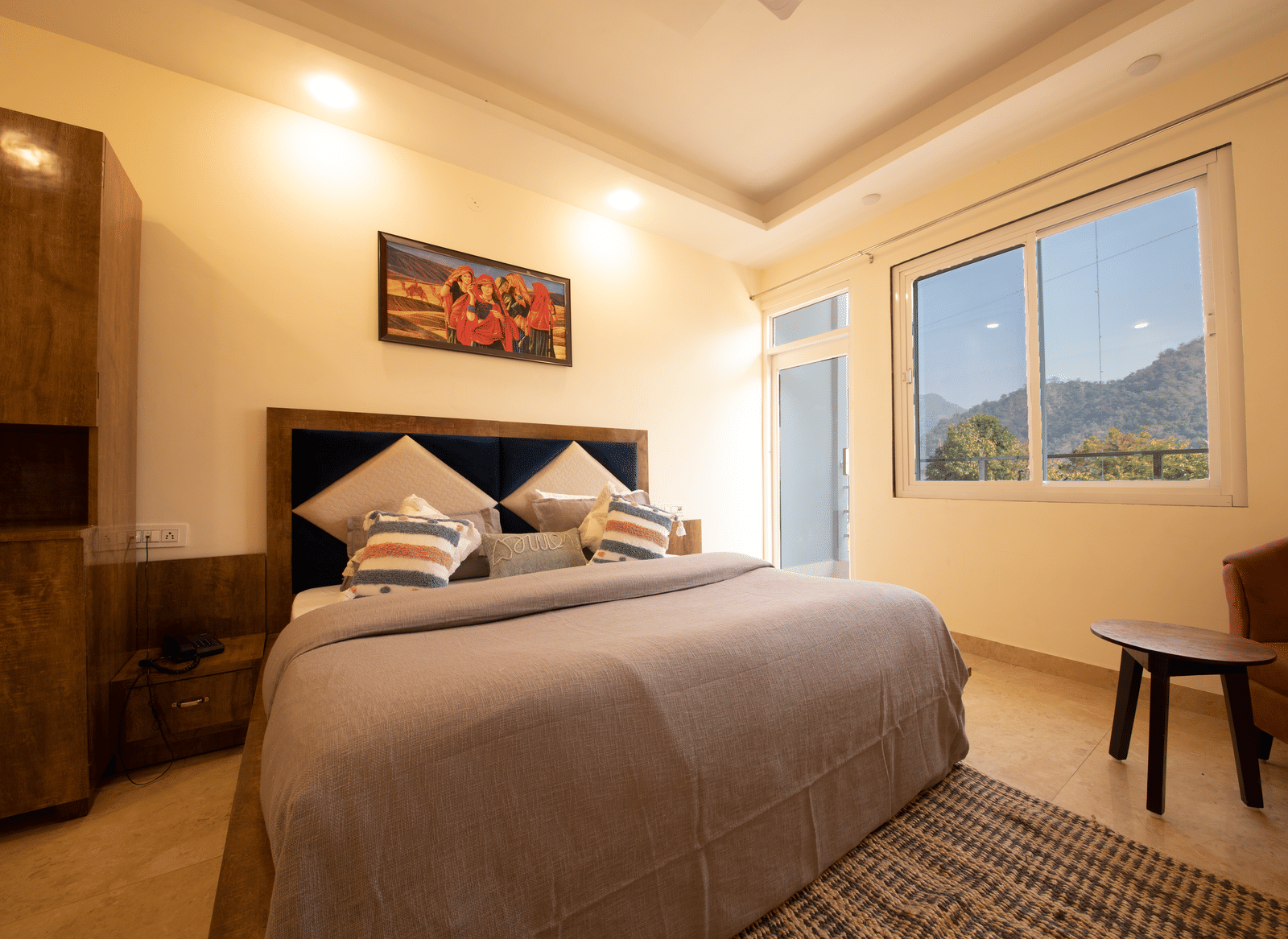 A hotel room, featuring a large bed with a grey duvet and a wooden frame, a painting on the wall, and a window with a view of hills | Perfectstayz Value Hills