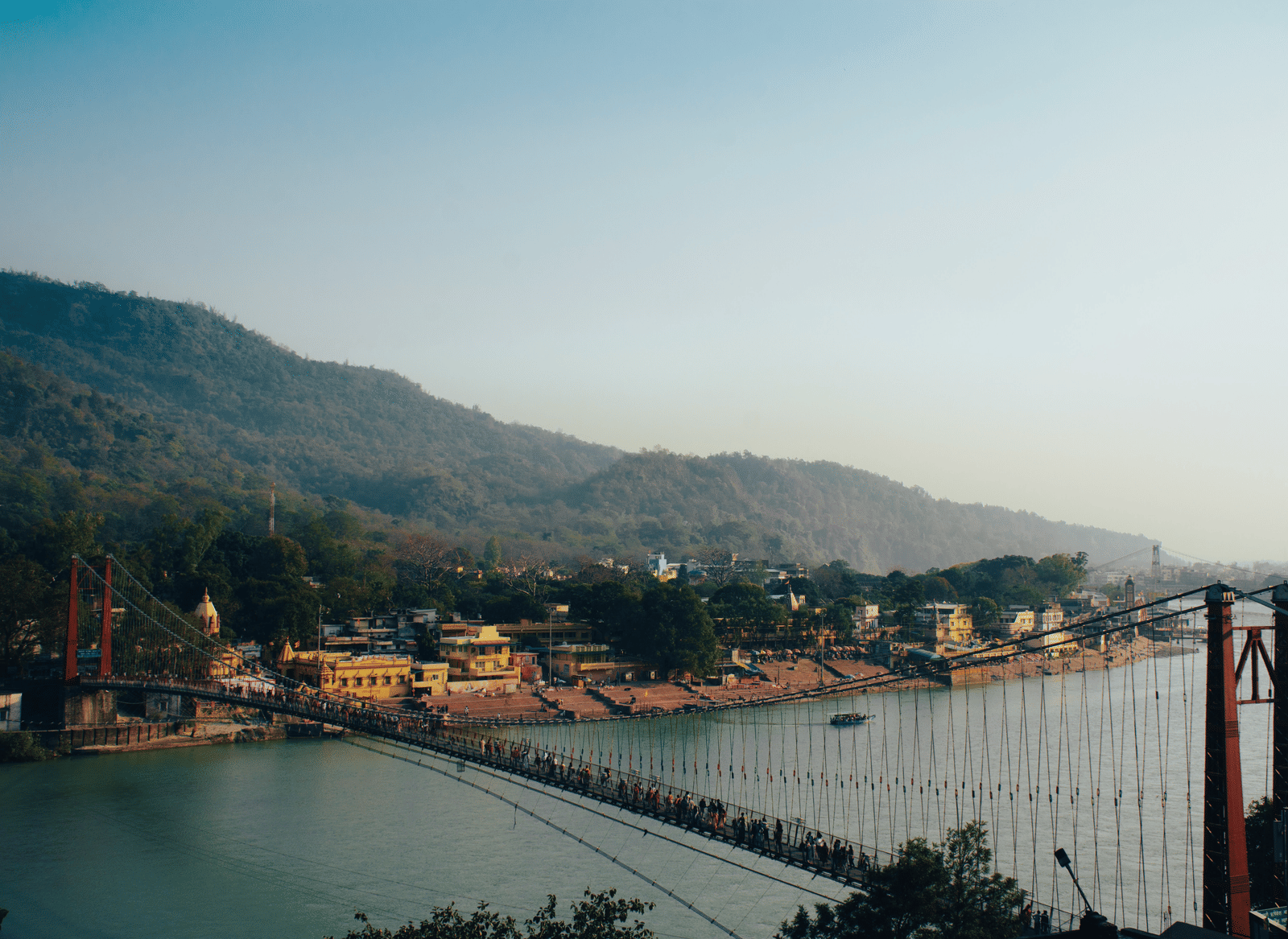 A long suspension bridge spanning a river in a valley, with hills and a town visible in the background