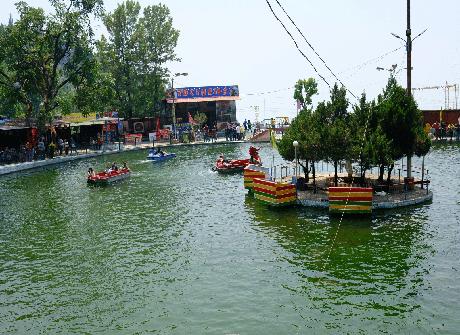 A boating lake with colourful paddle boats and a small tree-covered island surrounded by visitors enjoying the water.