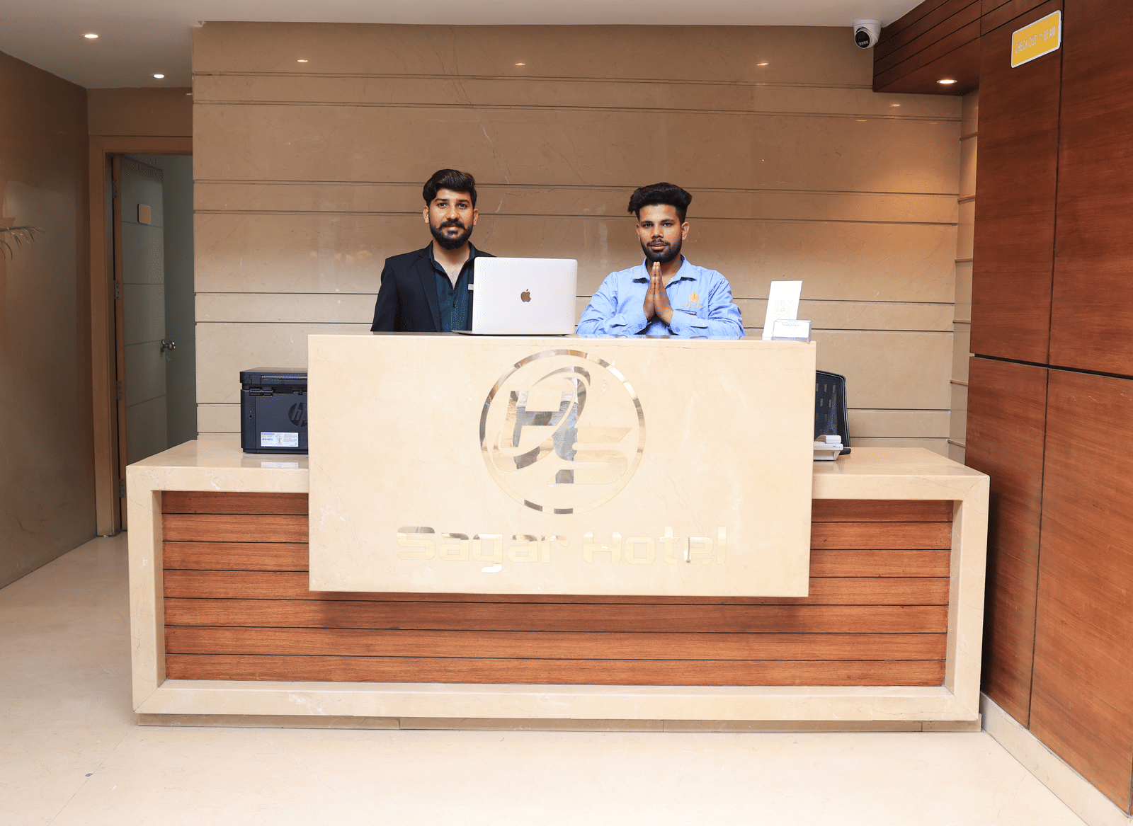 A hotel reception desk made of wood and light stone, with 2 male staff members standing behind it | Perfectstayz Value Sagar