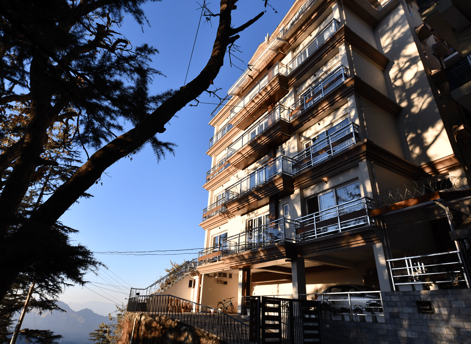 A facade of Perfectstayz Value Shimla (Namah Retreat) with balconies viewed from below against a clear sky and a tree in foreground.