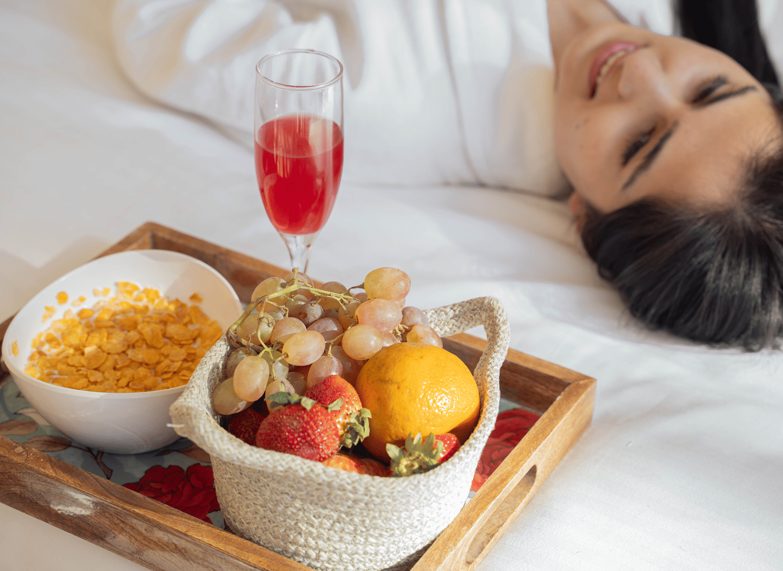 A tray on a bed with a bowl of cornflakes, a basket of fruits and a glass of red beverage, with a person lying beside it at Perfectstayz Value Shimla (Namah Retreat).