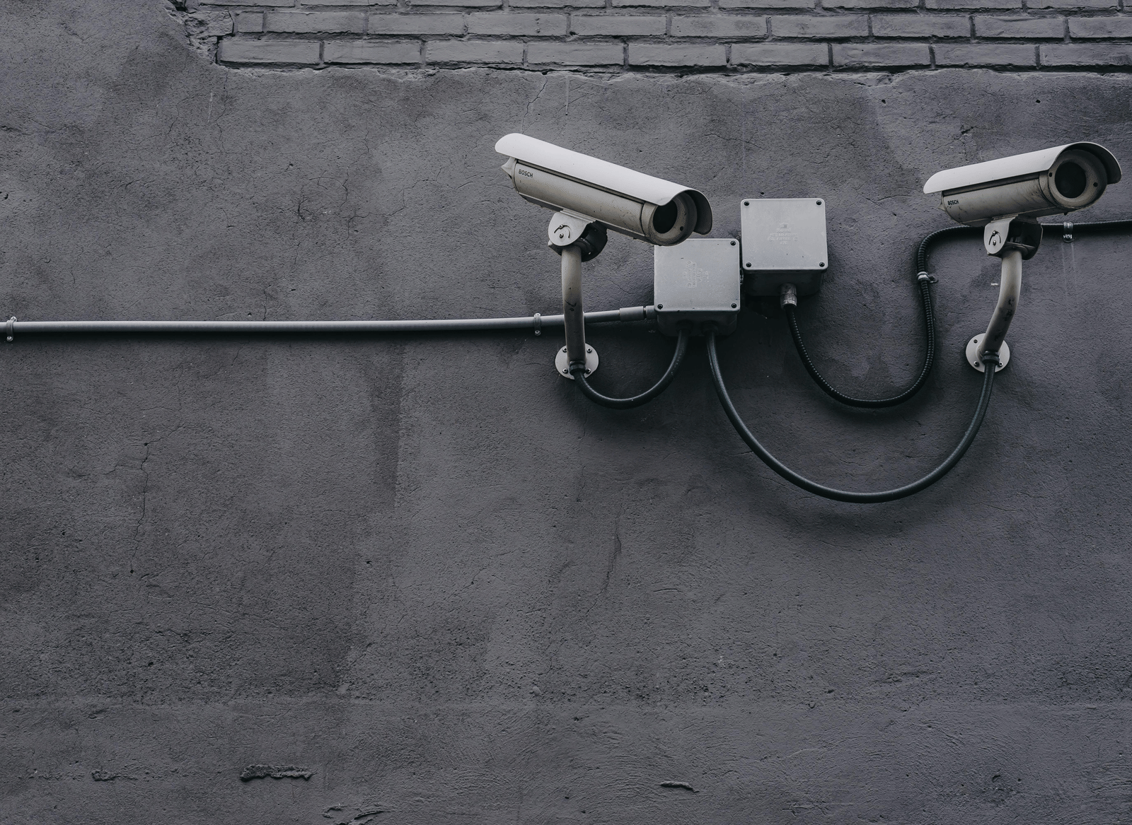 Two surveillance cameras mounted on a grey concrete or brick wall, with visible wiring.