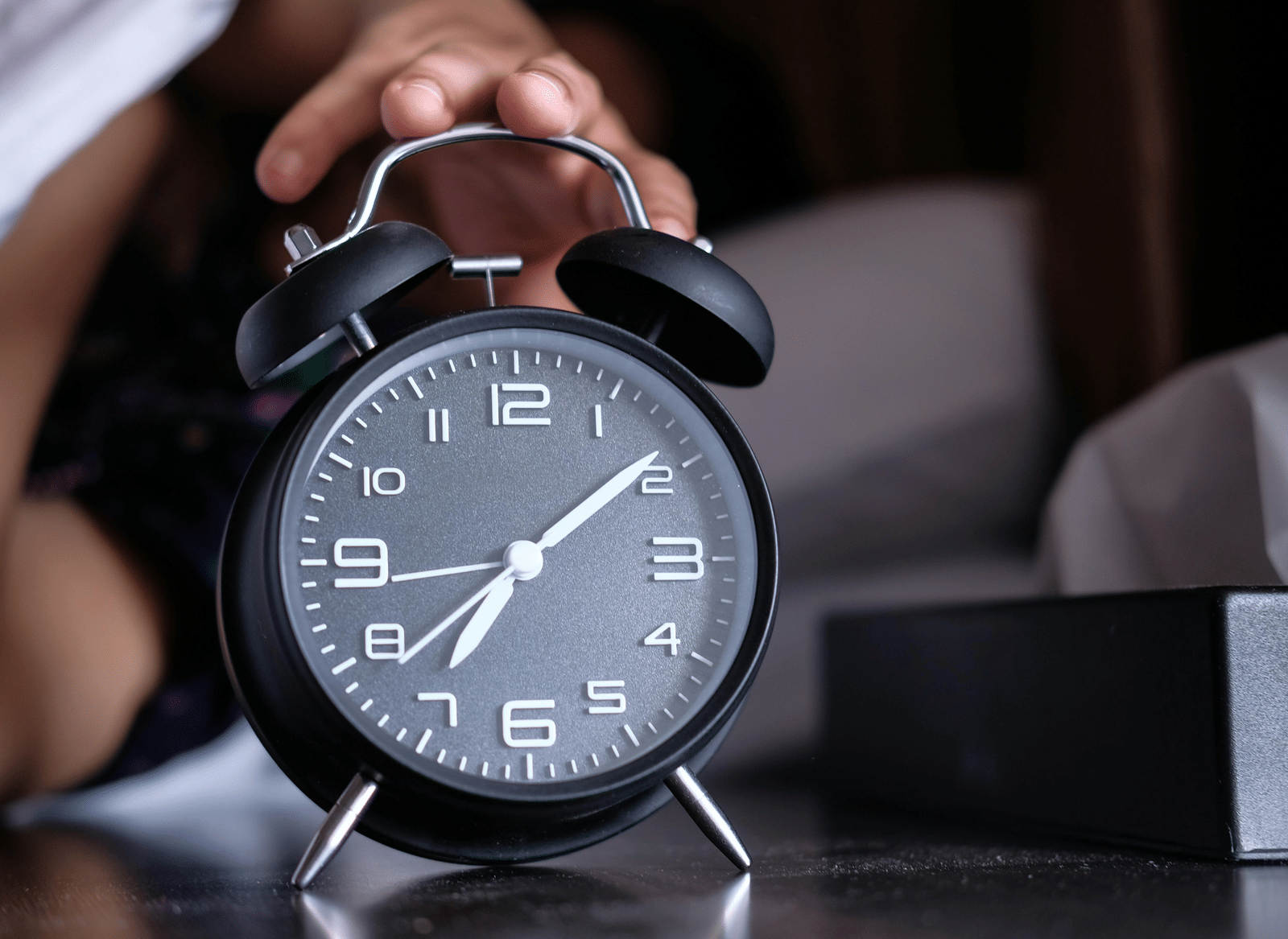 A hand reaching to silence a retro black alarm clock showing the time 7:00, next to a bed.