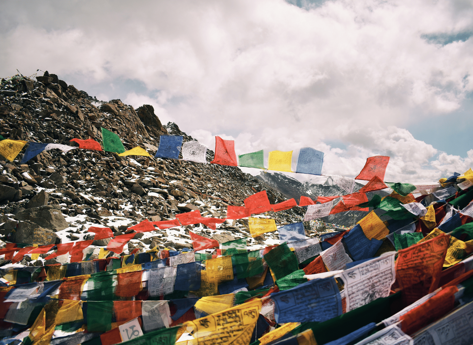 A mountain landscape with a rocky peak, covered in colourful Buddhist prayer flags waving under a cloudy sky