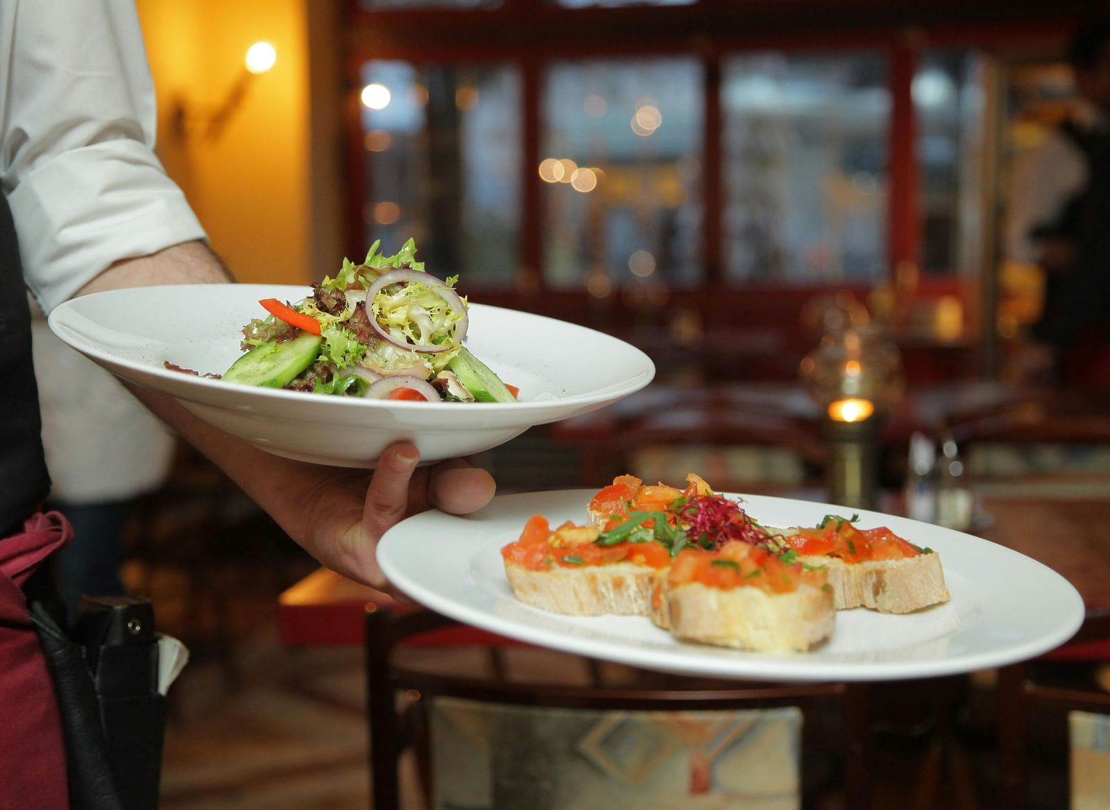 A person in a restaurant holding two plates with delicious dishes in it that are well plated.