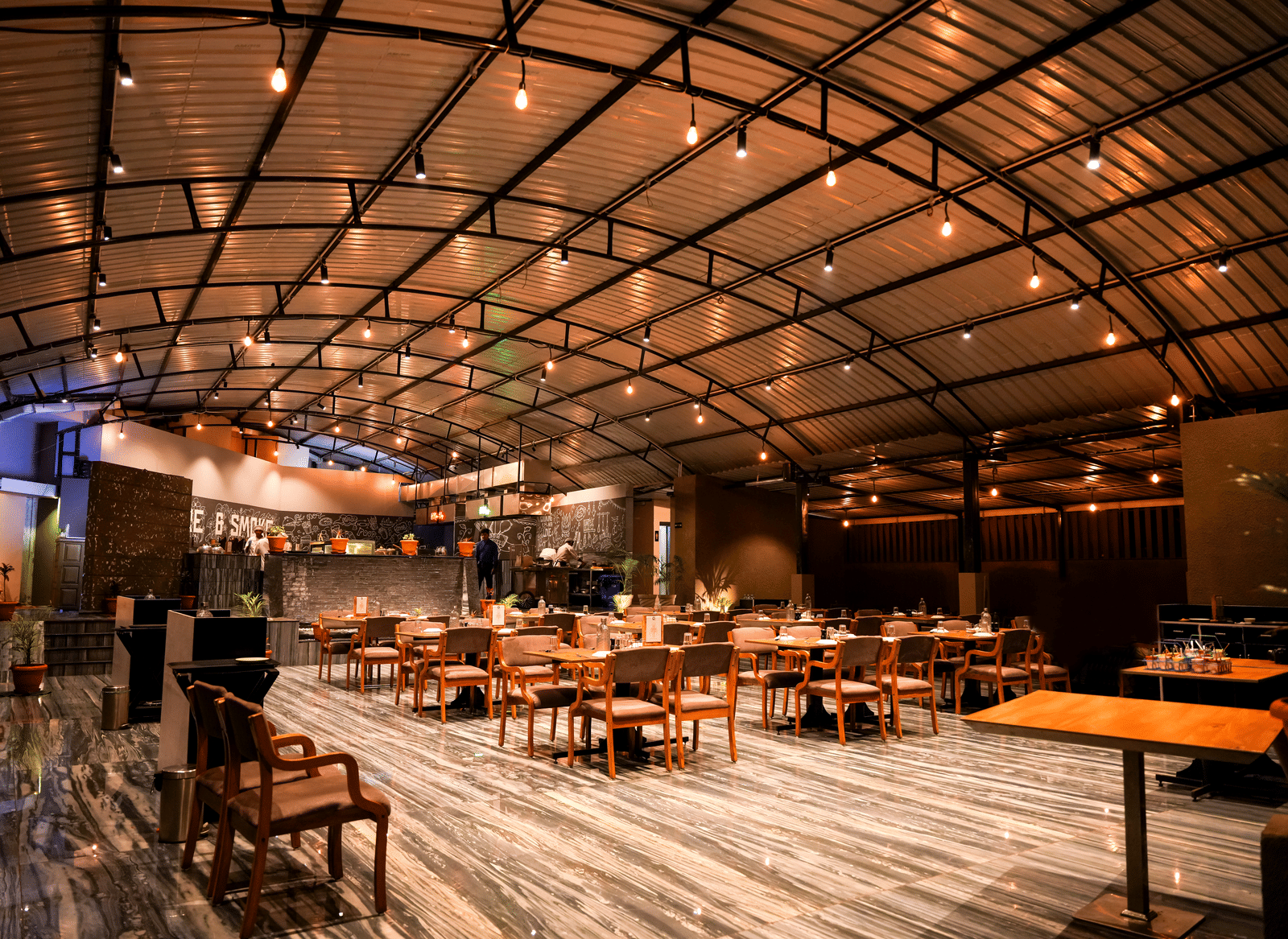  A wide view of the spacious dining hall with wooden tables and chairs under a curved, industrial-style roof at Pravasa Hotel, Kolhapur.