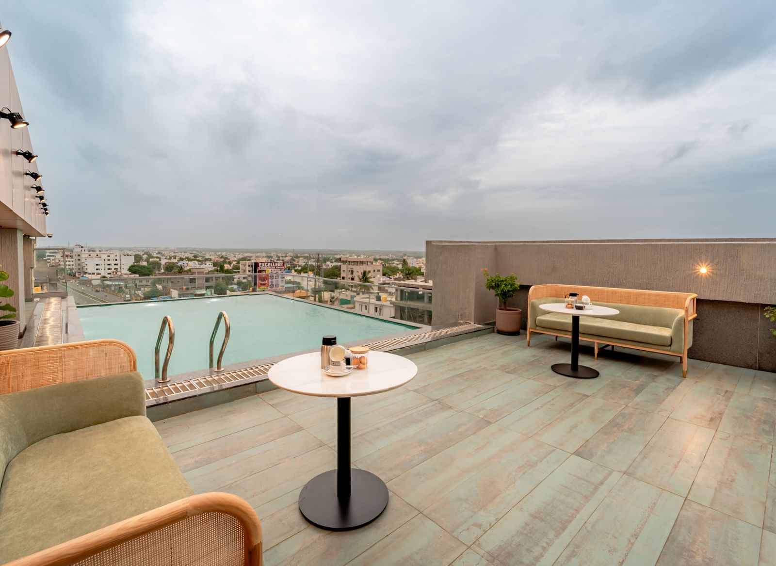 The rooftop swimming pool with clear water, surrounded by a tiled deck and some modern outdoor seating, offering a panoramic view of a city under a cloudy sky | Pravasa Hotel Vijayapura