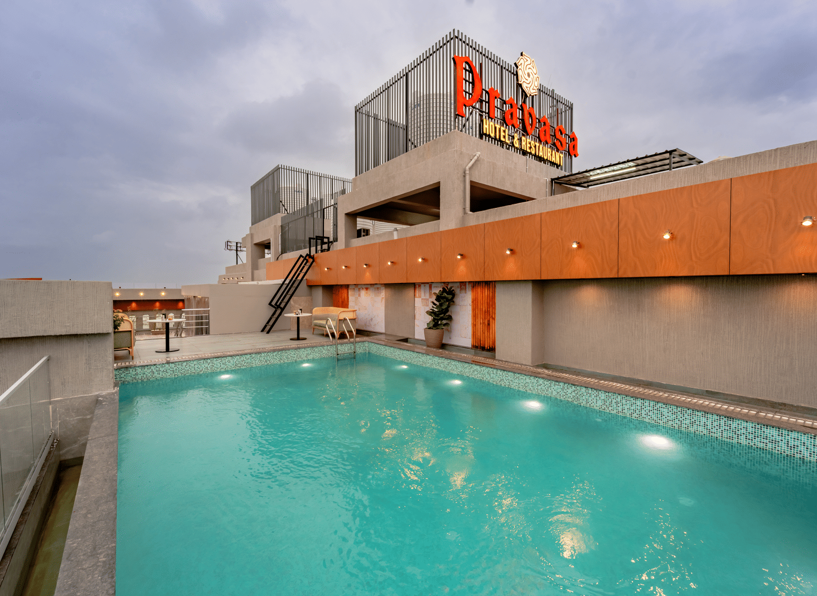 A rooftop swimming pool surrounded by a tiled deck and some modern outdoor seating, offering a panoramic view of a city under a cloudy sky | Pravasa Hotel Vijaypura
