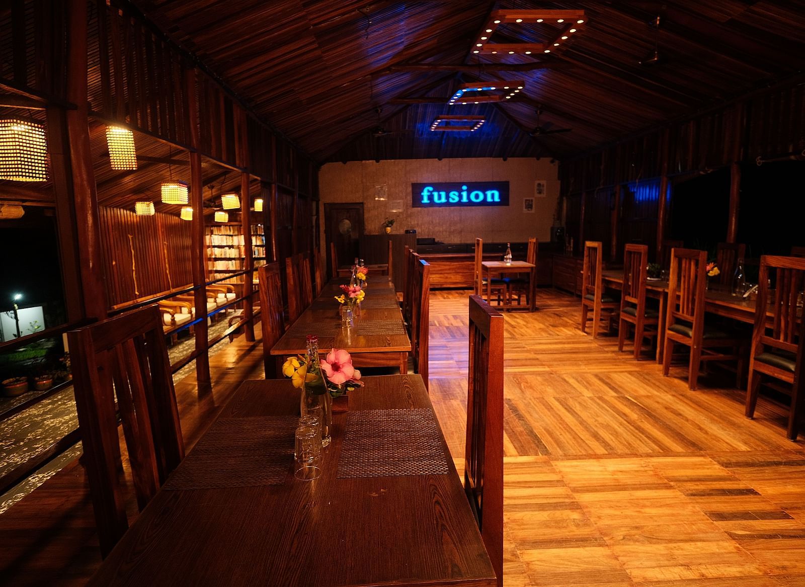 A wooden interior is illuminated by hanging lamps, highlighting the seating and shelving arrangements | Fusion Cafe and Multi-cuisine | RainForest Resort, Port Blair, Andaman and Nicobar Island