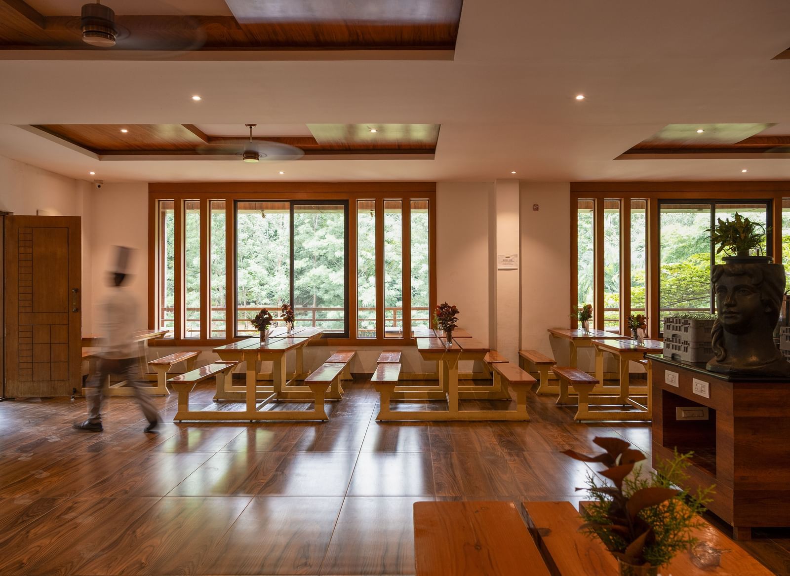 A spacious dining hall with wooden floors and high ceilings offers multiple tables and chairs, with large glass doors opening to an outdoor area | Lake View Restaurant