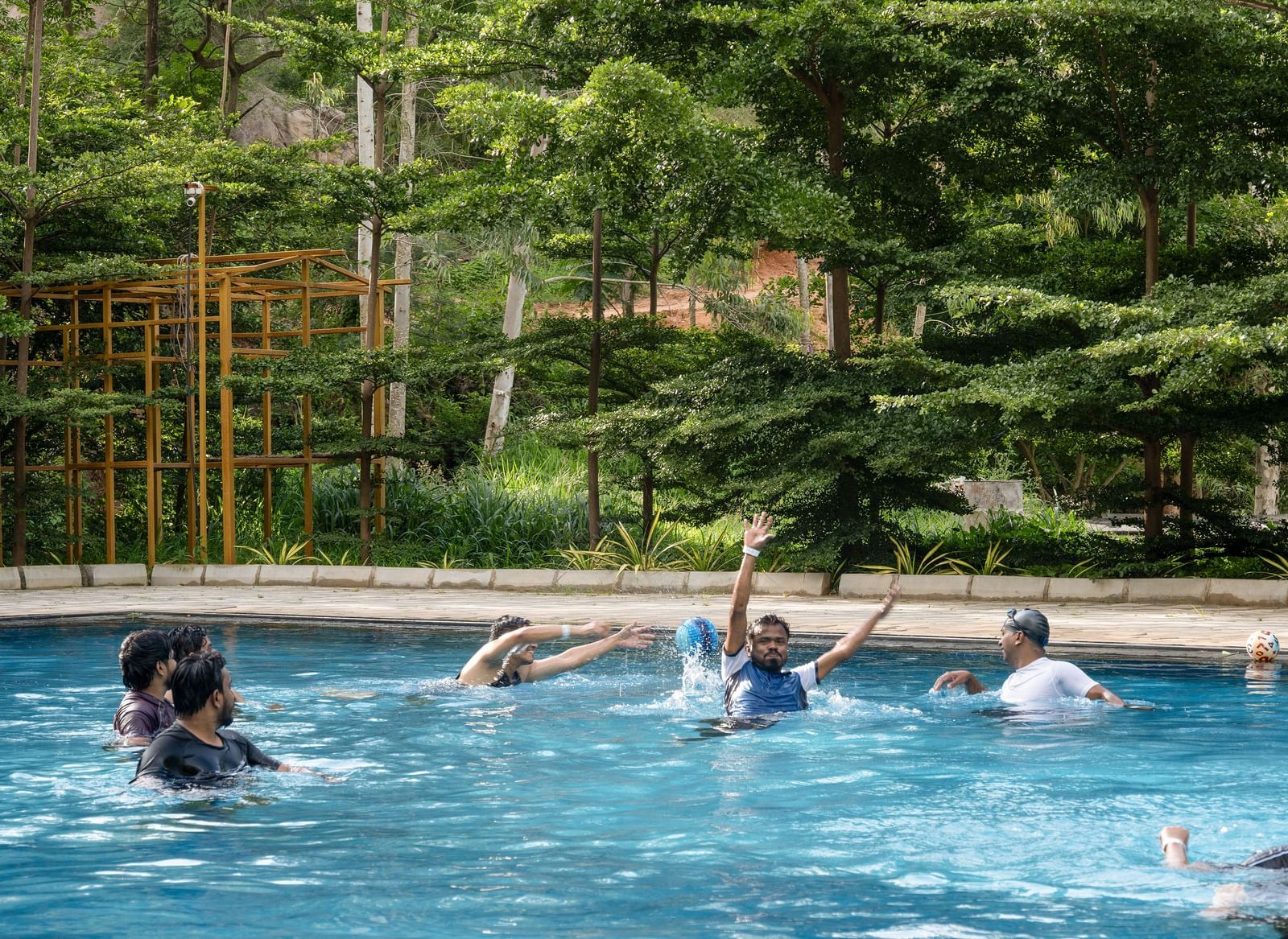 Several people are enjoying a swimming pool surrounded by tall trees and lush vegetation, indicating a natural or resort-like setting | RD's Nature Retreat, Bangalore