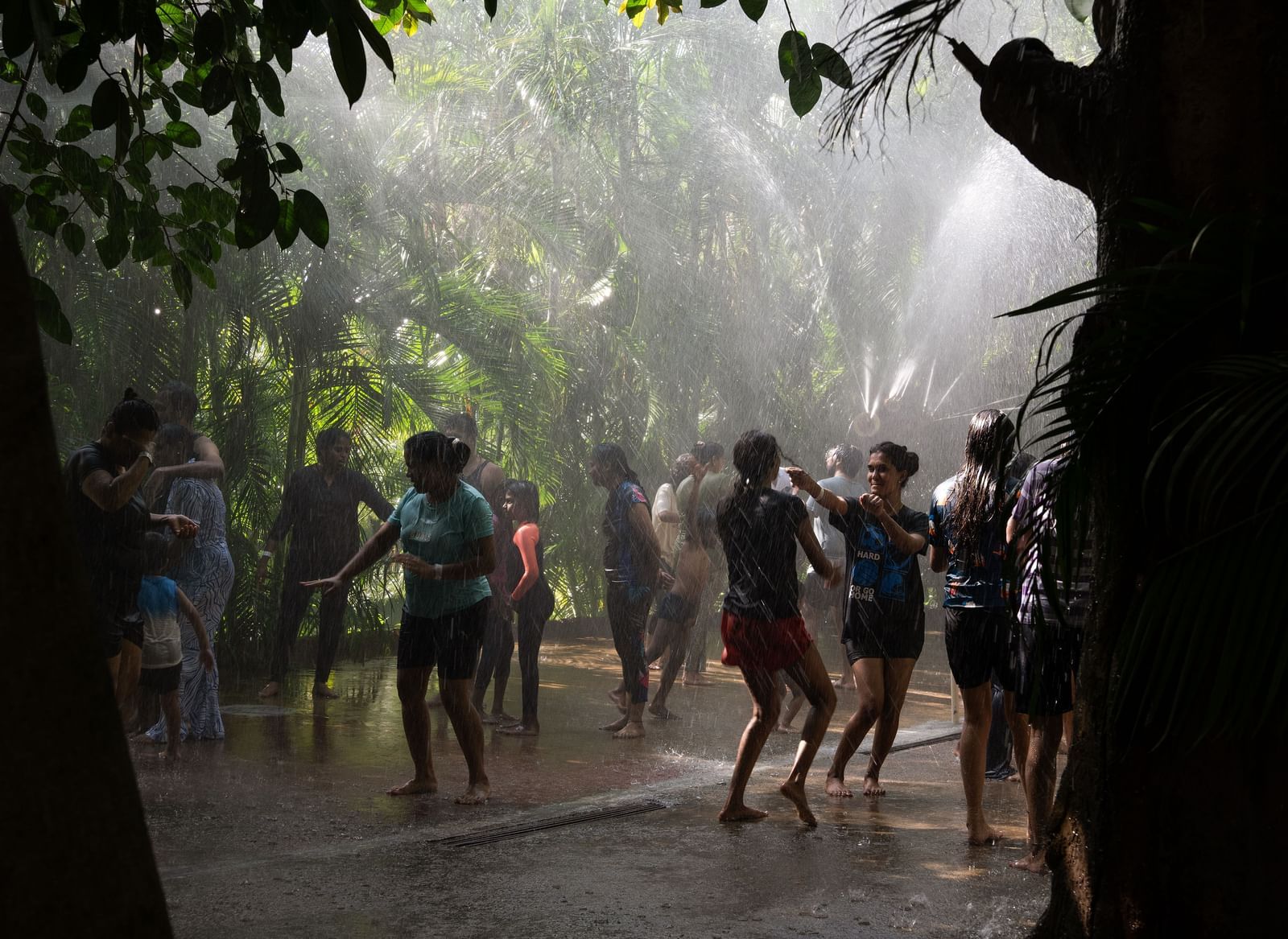 A group of people are seen enjoying a rain dance or water activity, with water spraying down on them in a shaded, outdoor setting | RD's Nature Retreat, Bangalore