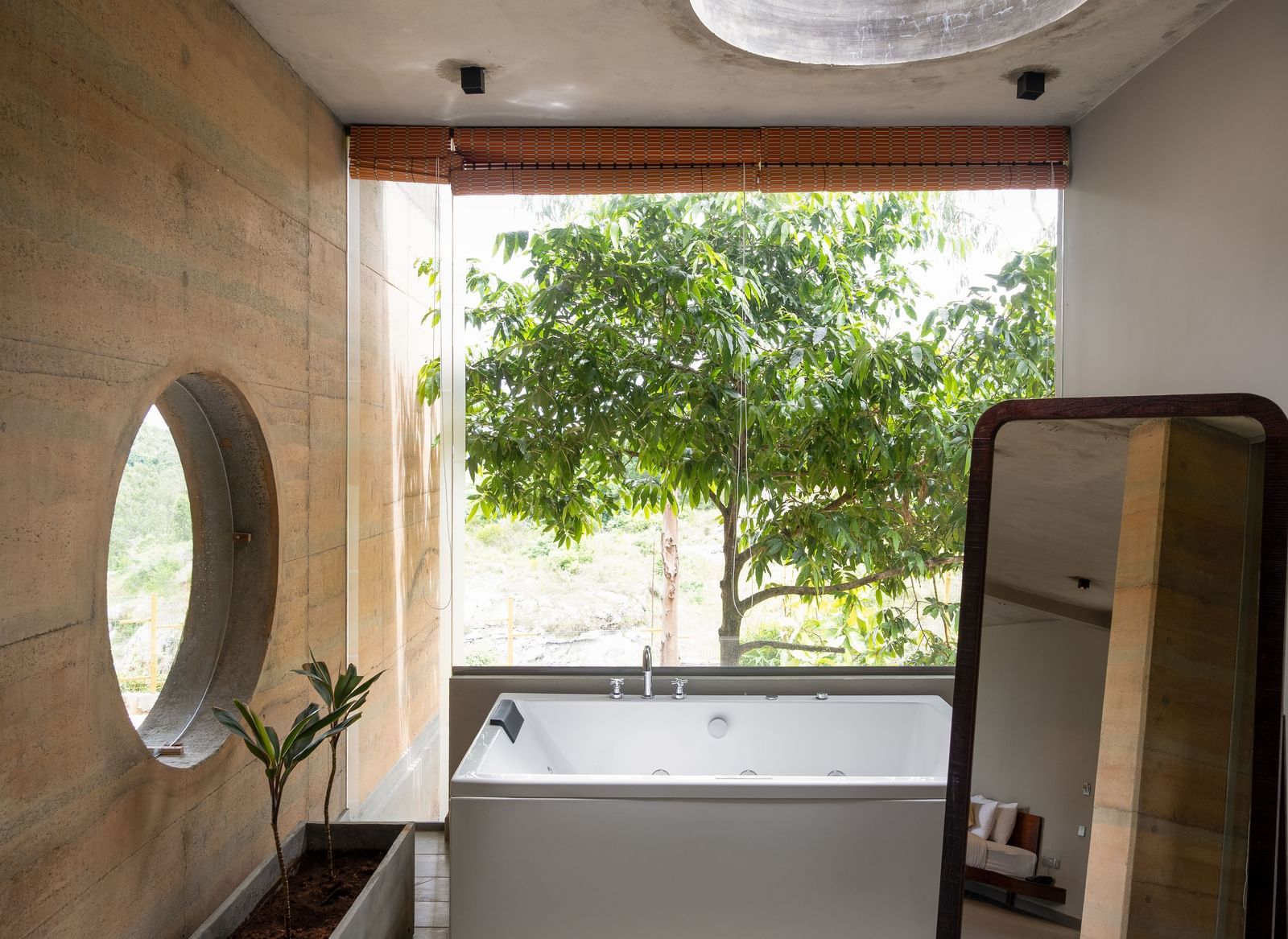 A bathroom featuring a large round mirror, a vanity area, and a window overlooking lush trees | Gaia Studio | RD's Nature Retreat, Bangalore