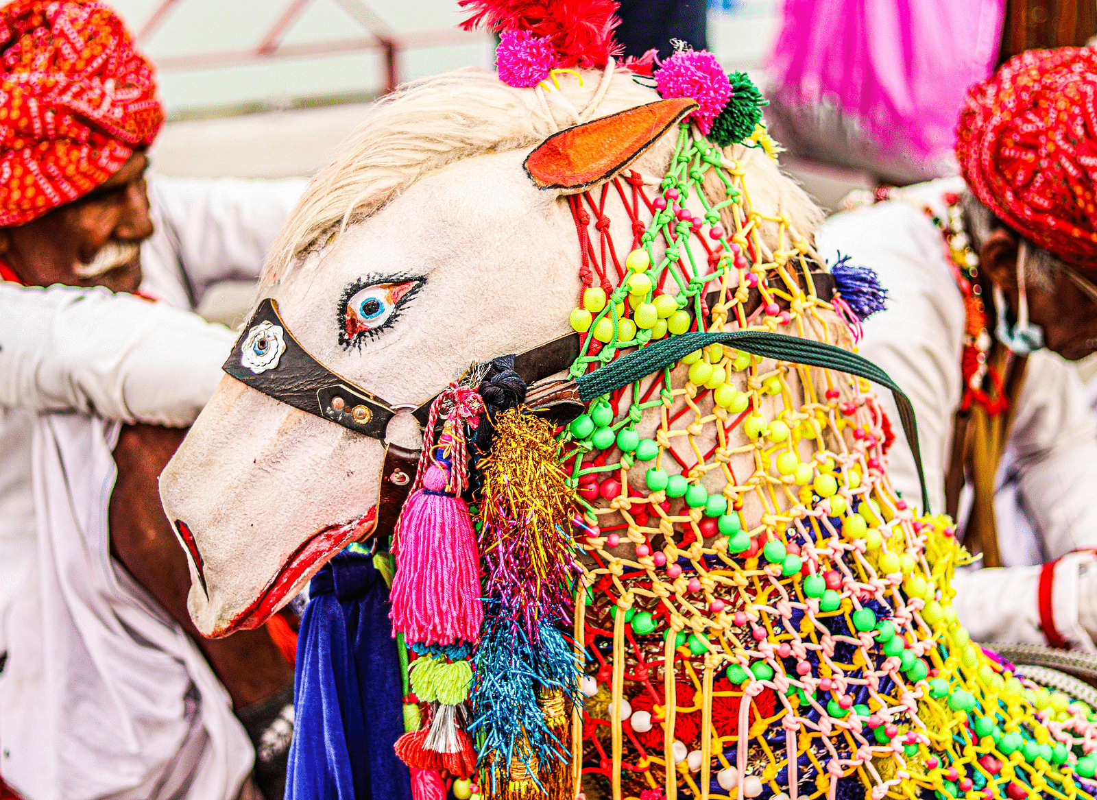 Two Rajasthani men in traditional attire, including colourful turbans, adjust a decorative, white horse-shaped puppet adorned with beads, tassels, and a saddle, preparing it for a cultural performance.