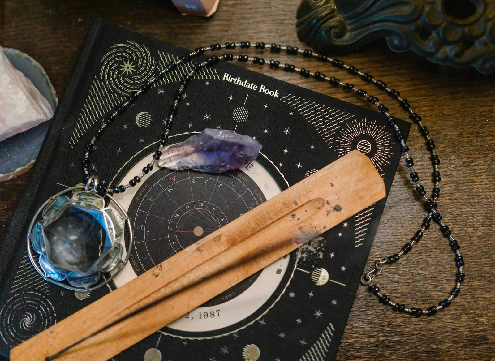 Astrology book with a celestial chart design, surrounded by a long black beaded necklace, incense, and a blue crystal.