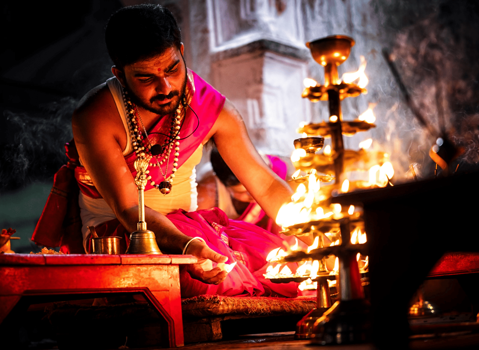 A priest in a pink dhoti and shawl, performs a fire ritual (Aarti or Havan), tending to multiple flames on a pedestal with multi-tiered brass oil lamps.