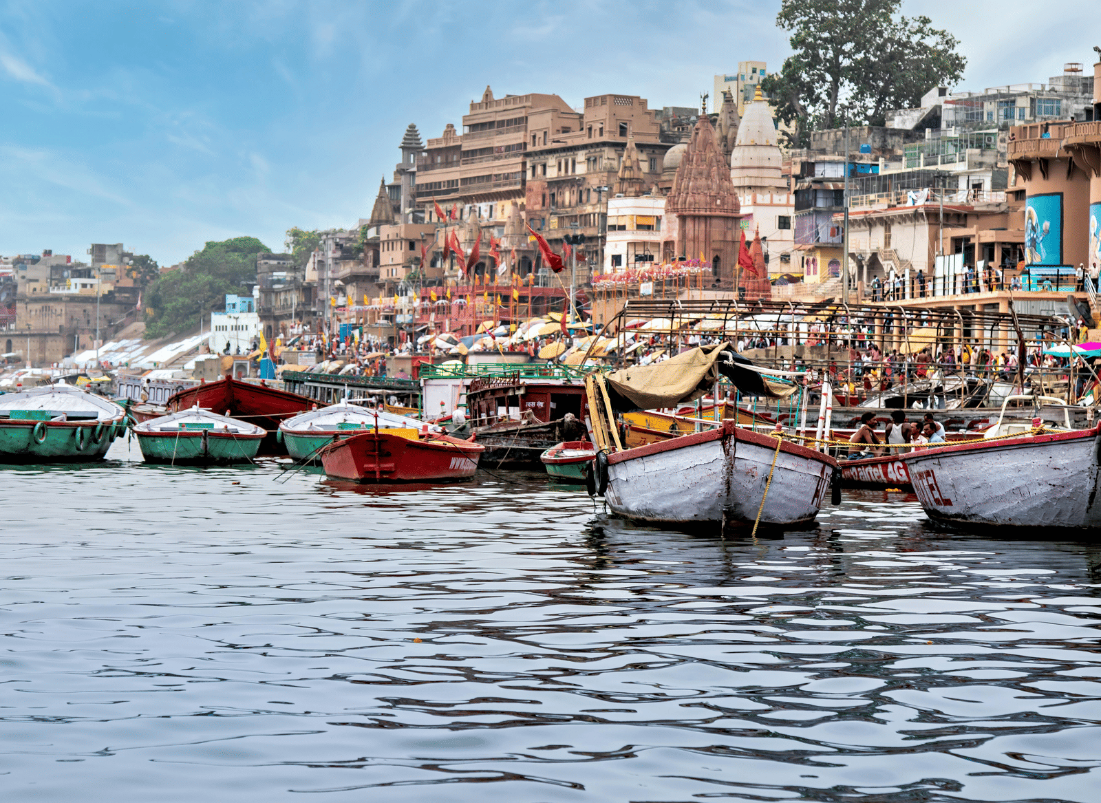 Small wooden boats moored on the Ganges River near the steps (ghats) of a crowded riverside city in India.