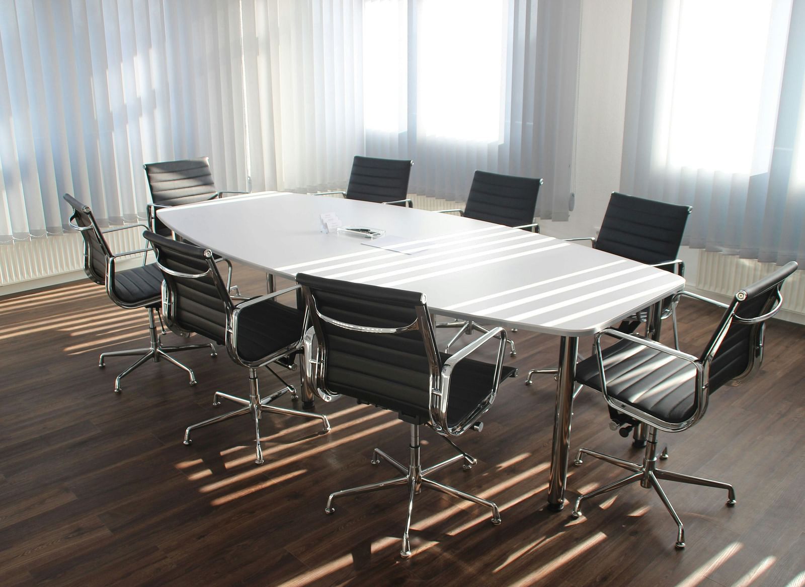 A close up shot of a brightly lit boardroom featuring a white long table along with office chairs surrounding it.