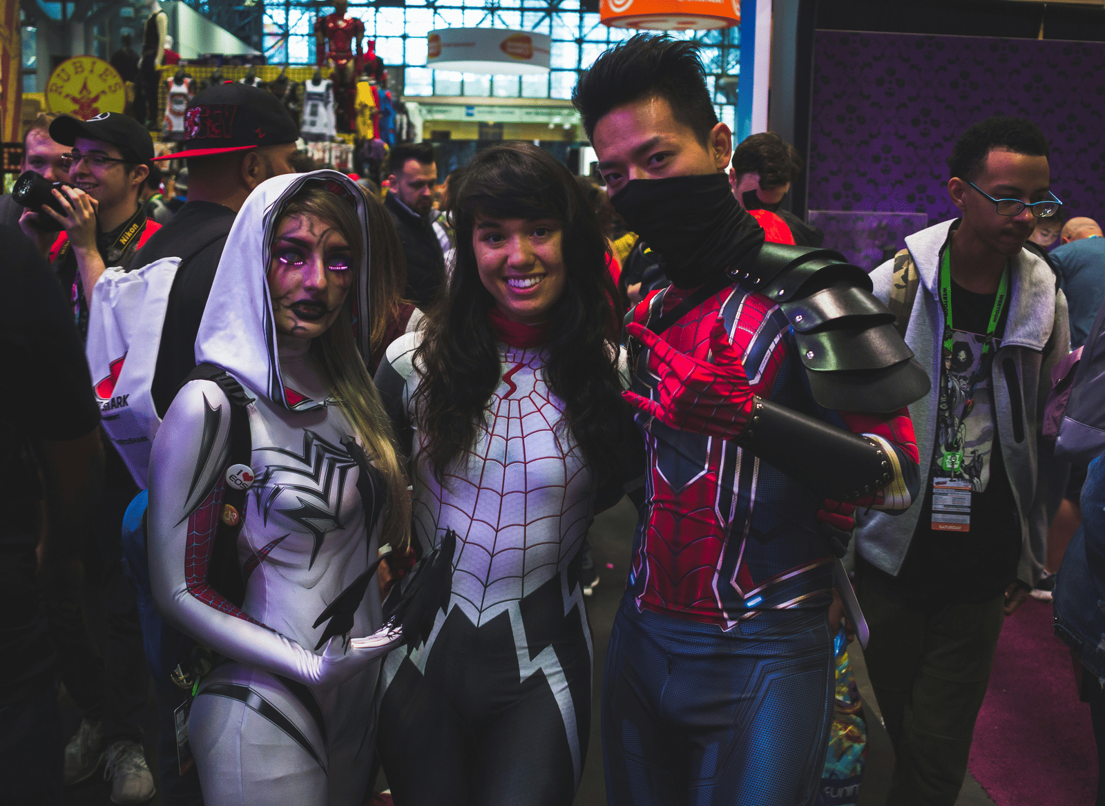 Three people in detailed superhero-themed cosplay posing together at a crowded convention.