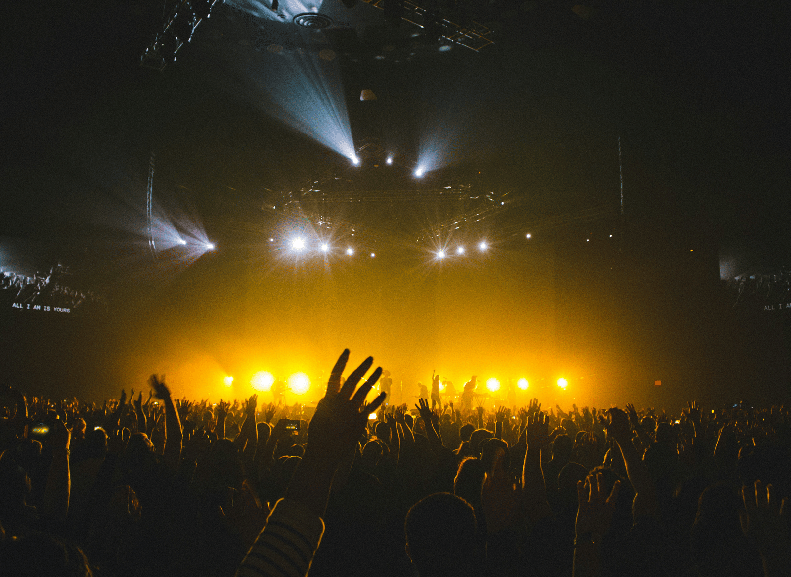 A large crowd at a concert raising their hands toward a brightly lit stage with intense yellow lighting and spotlights overhead.