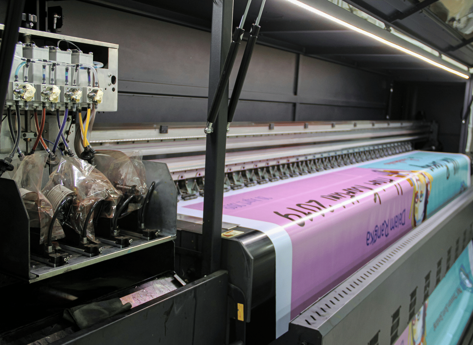 A large-format printer is printing a colourful banner, with visible ink supply tubes and part of the design in purple and blue, showing text and graphics.