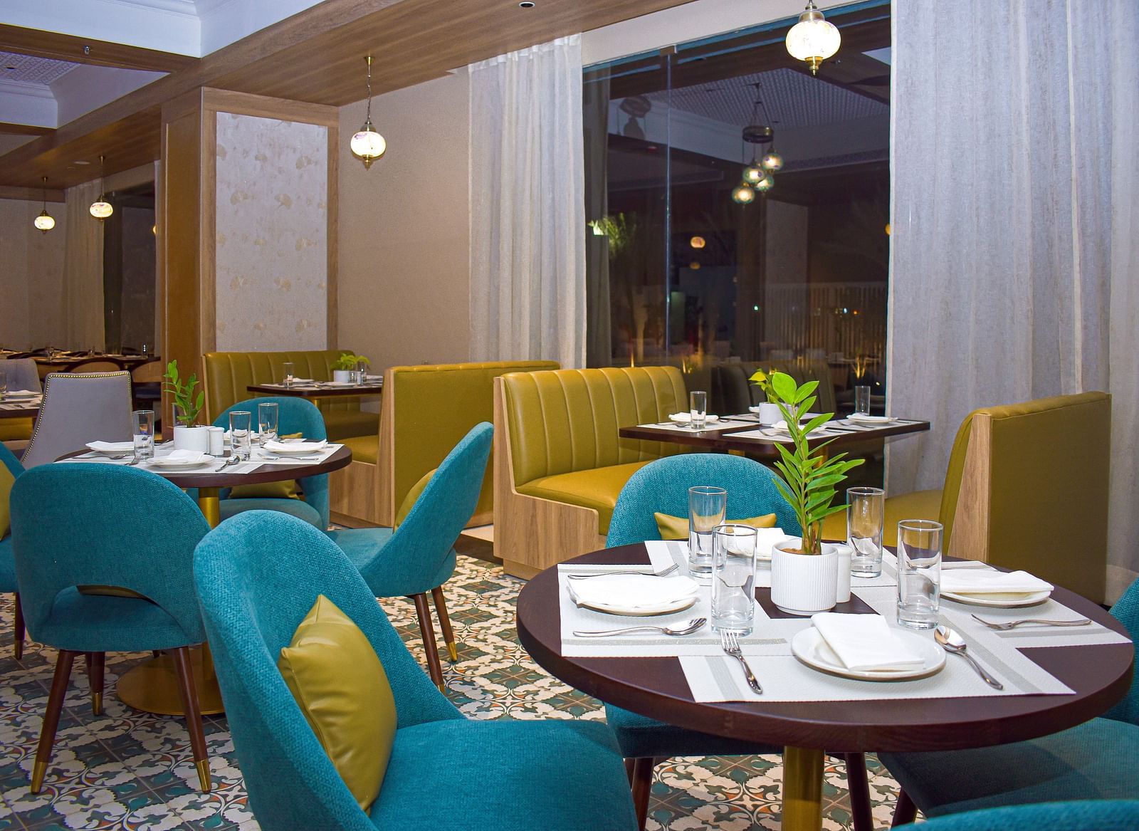 A restaurant interior with teal armchairs, booth seating, round tables, patterned floor, sheer curtains, and hanging pendant lights - Urban Suites by BluSalzz, Jaipur