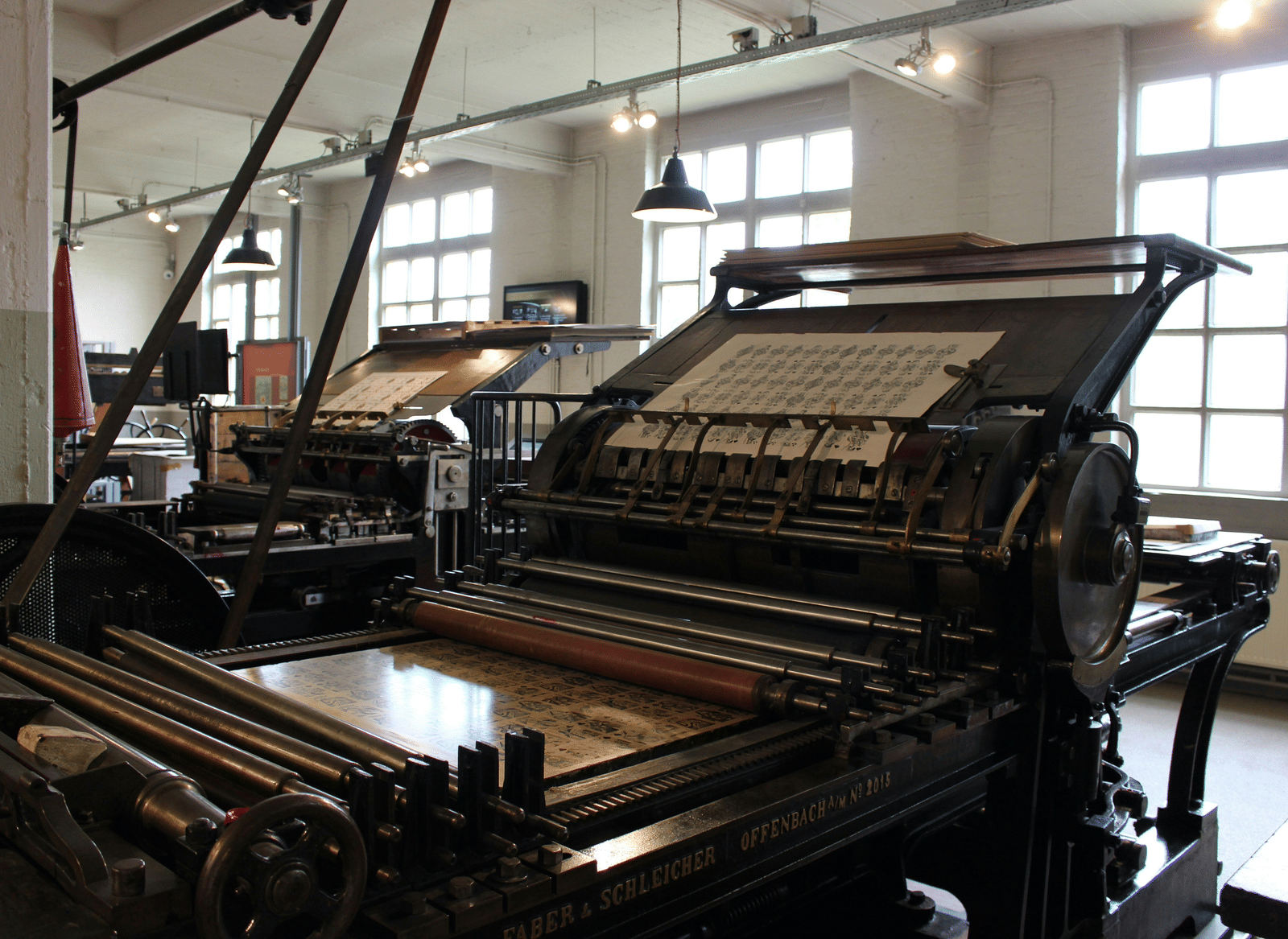 A room filled with large vintage printing presses, showing metal rollers, gears, and printed sheets, illuminated by overhead lights and large grid windows.