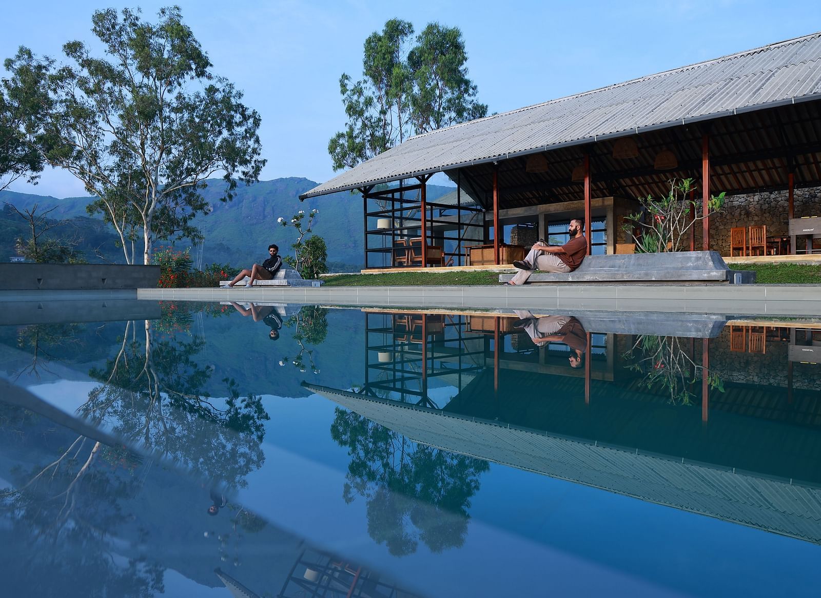 Two people relax by an infinity pool at Vaasta Vagamon, overlooking a serene landscape with rolling hills and trees.