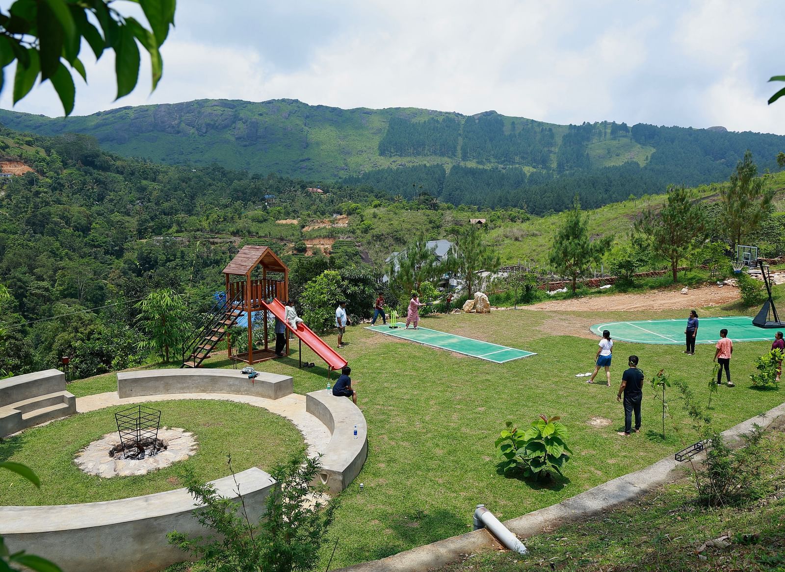 A vibrant outdoor recreational area at Vaastha Vagamon with a playground, sports court, and fire pit, surrounded by lush hills.