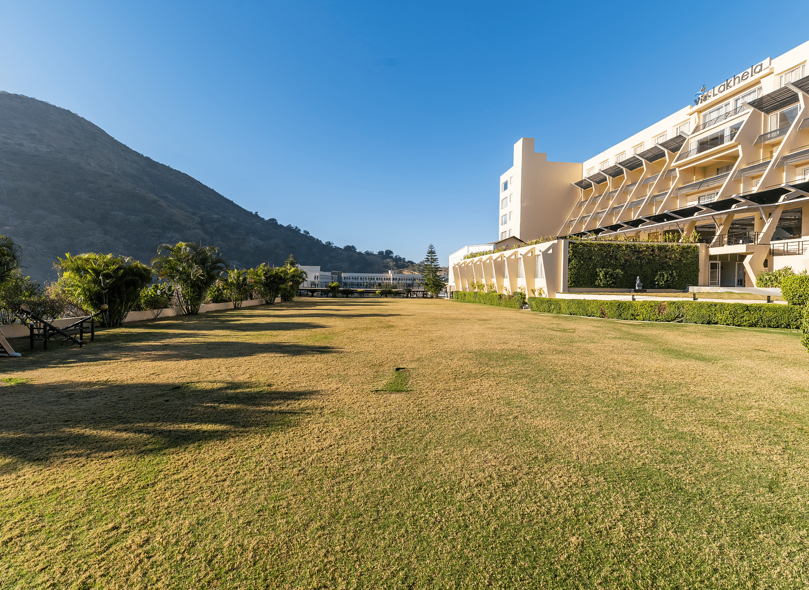 A wide shot of a building with a mountain backdrop in a green field - Via Lakhela Resort & Spa