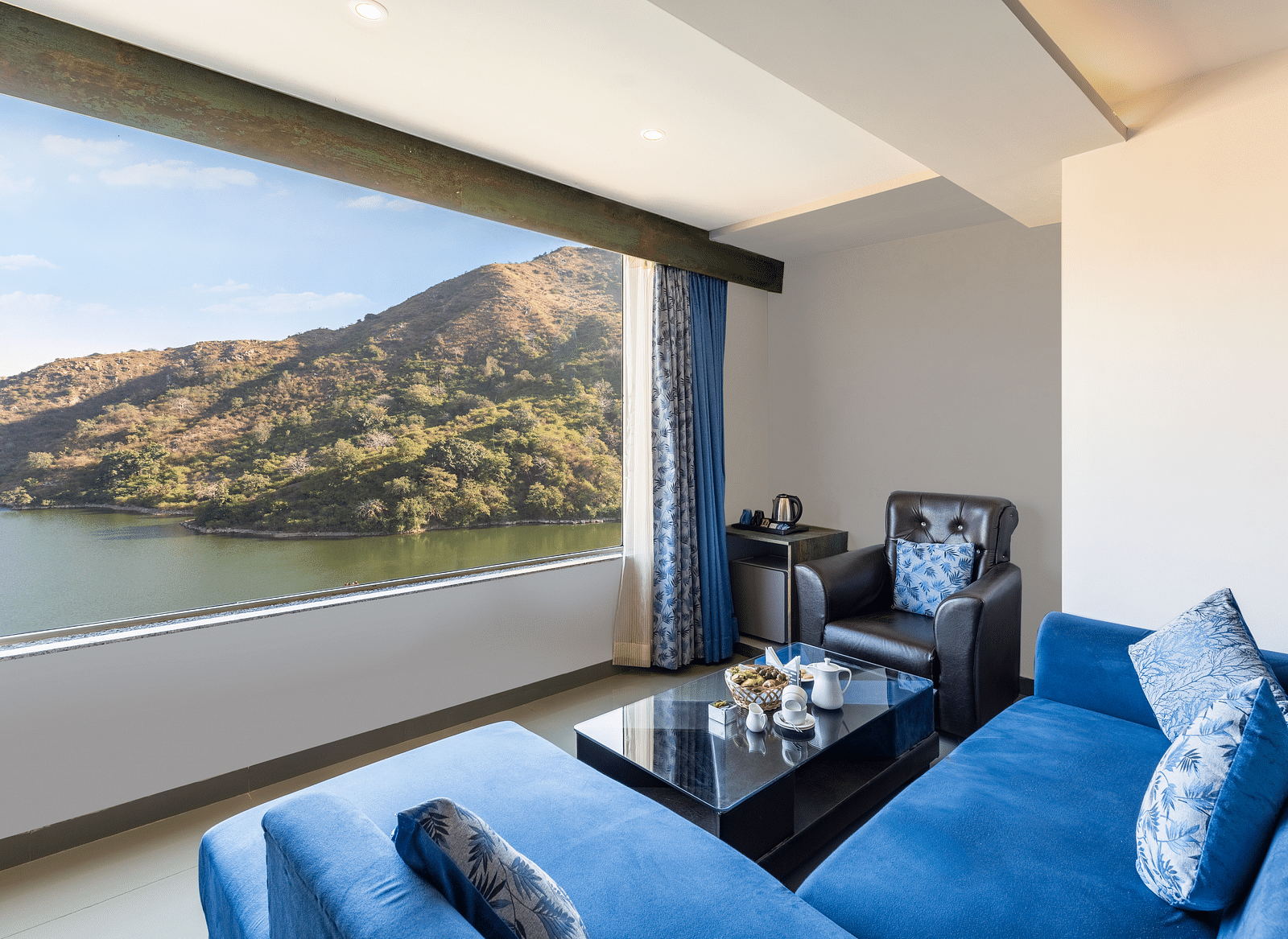 Living area with a blue couch and a large window with a view of mountain and water - Via Lakhela Resort & Spa