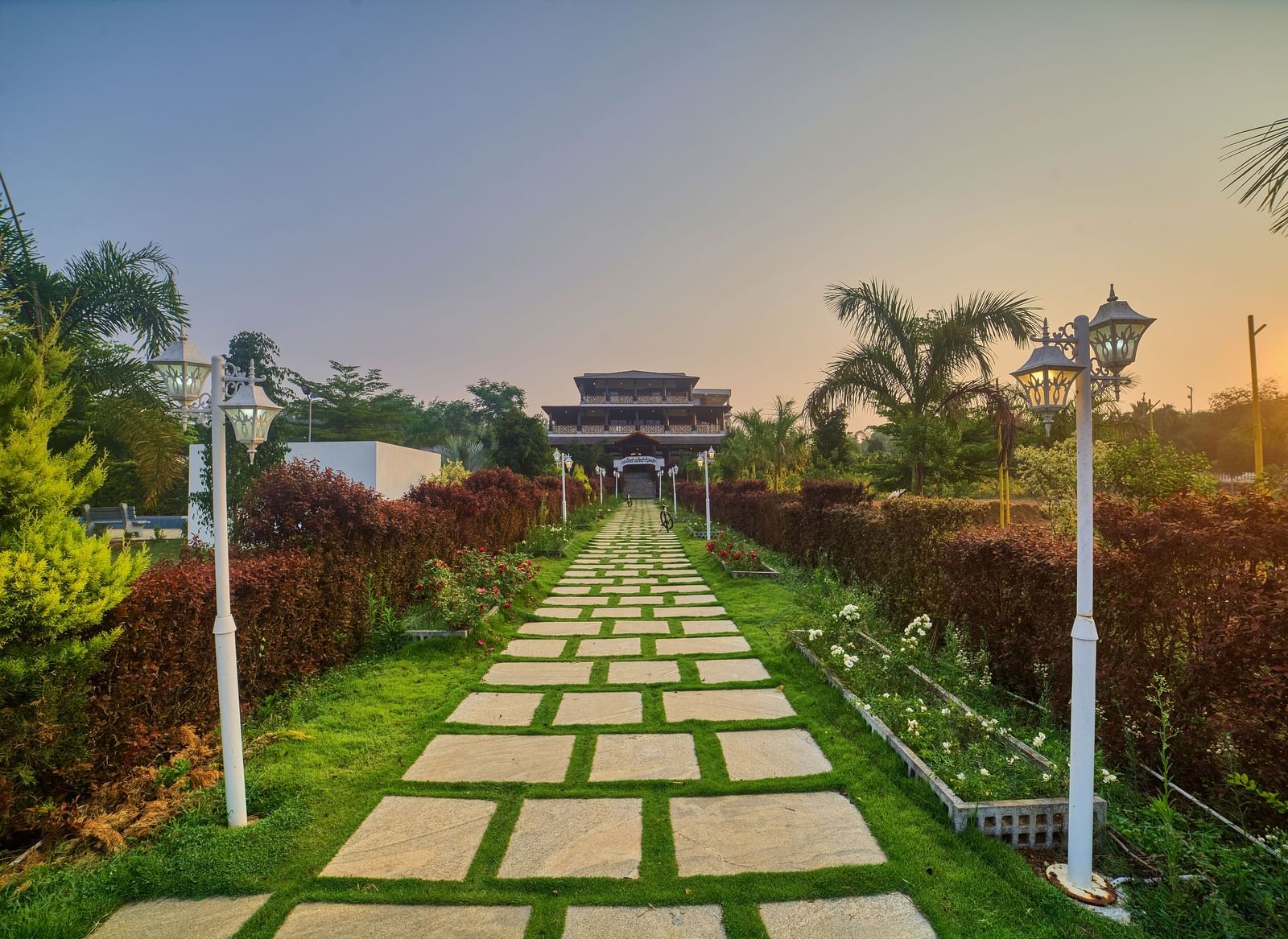 A long pathway made of square stepping stones through a manicured green lawn, leading towards a distant entrance structure or building under a sunset sky. - Vintage Trails by Ilara