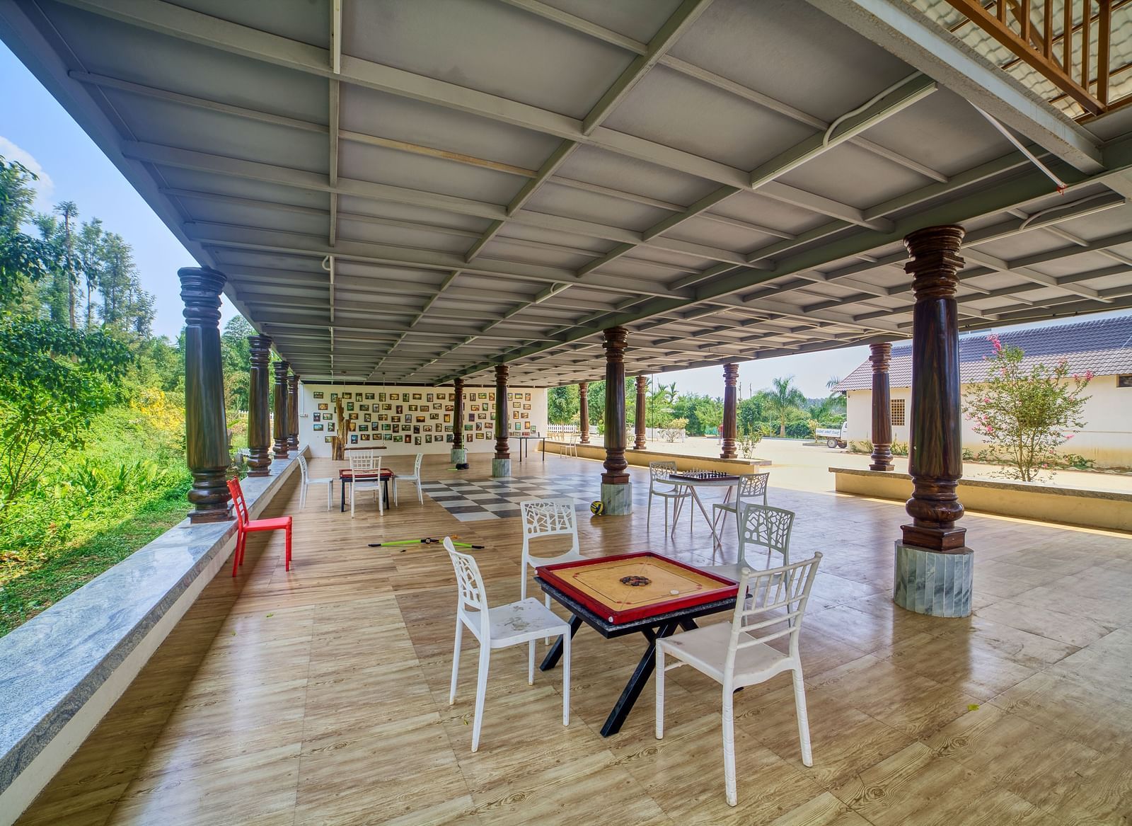 A spacious, covered outdoor area with a dark tiled floor, featuring a carrom board table and white chairs, overlooking a green landscape. - Vintage Trails by Ilara