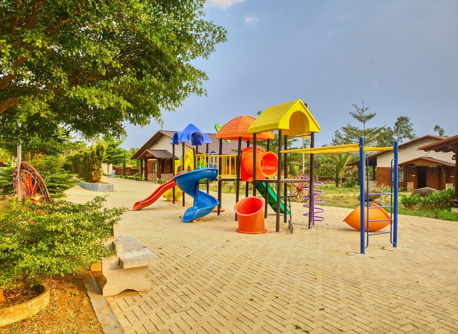 A colourful outdoor children's playground with various slides, climbing frames, and swings on a paved area, surrounded by trees. - Vintage Trails by Ilara