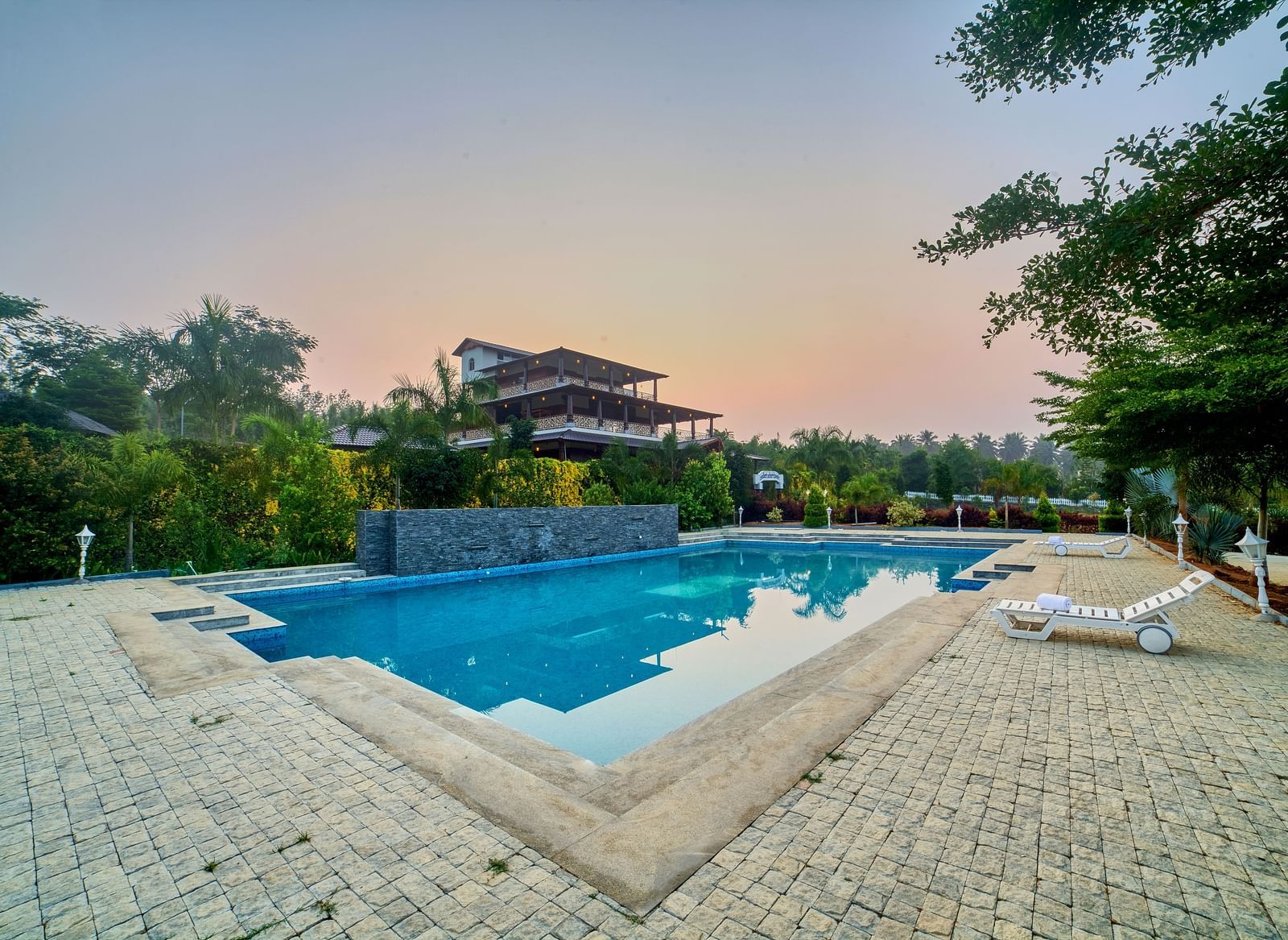 An outdoor swimming pool with a unique, geometric shape, surrounded by light-coloured paving stones and lush green trees, under a clear sky. - Vintage Trails by Ilara