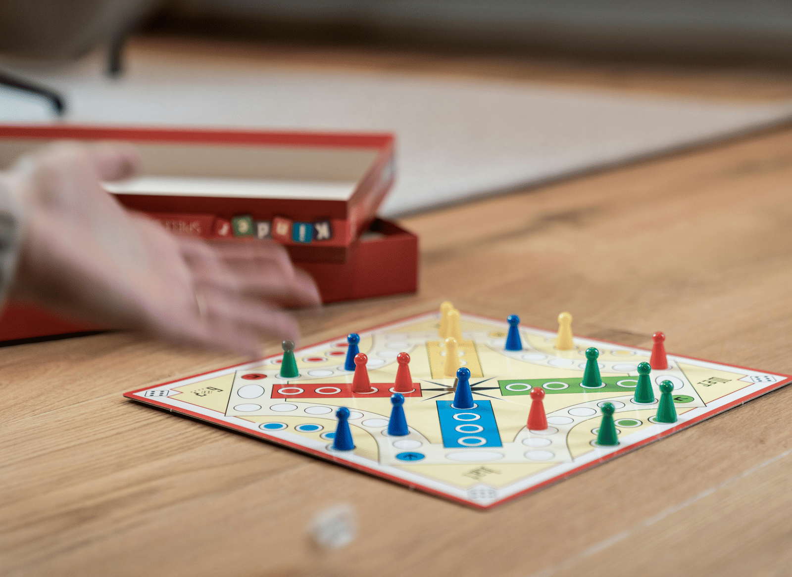 Two hands are playing a board game with red, green, blue, and yellow playing pieces on a wooden table.
