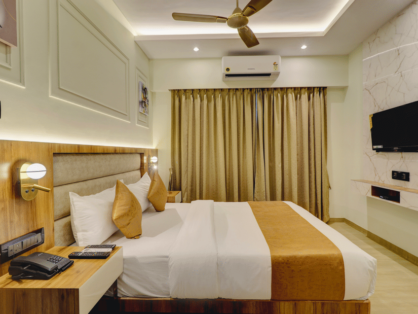 A bedroom at Home2 India with a king size bed, pillows, bedside table, wall-mounted TV, air conditioner, curtains, ceiling fan, and soft lighting.