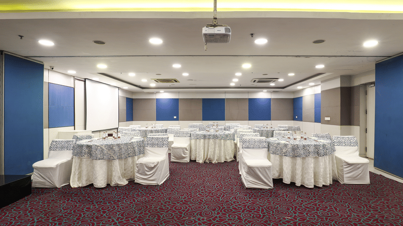A large and well-lit conference hall with round tables, white chair covers, and a modern projector setup, perfect for professional events and meetings.