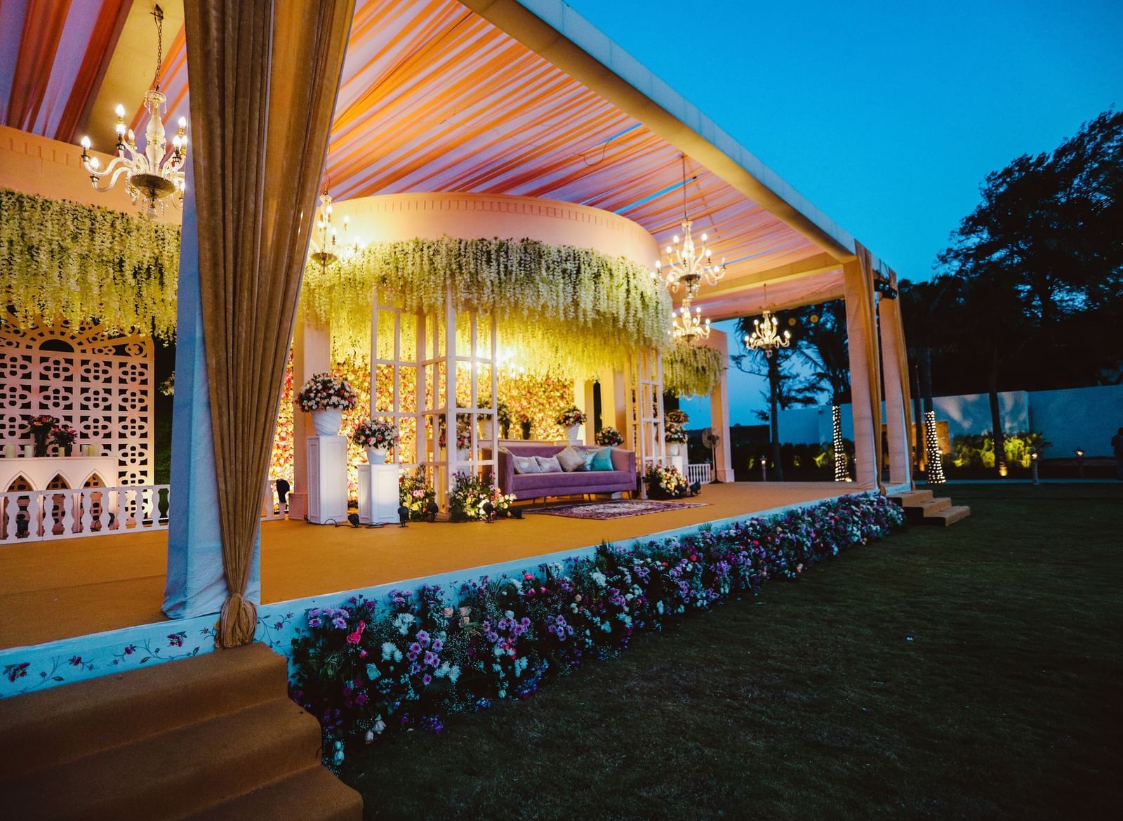 A stage decorated with flowers at the front set in a lawn, featuring ornamental plants, a chic sofa and chandeliers hanging from ceiling - Kaldan Samudhra Palace